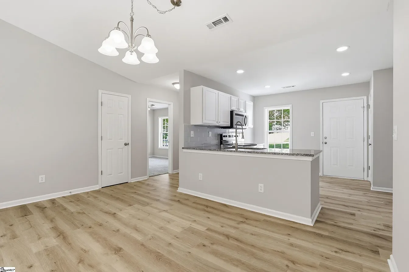 Empty living area with light wood flooring, white walls, a kitchen with white cabinets, granite countertops, stainless steel appliances, a window, and nearby rooms.