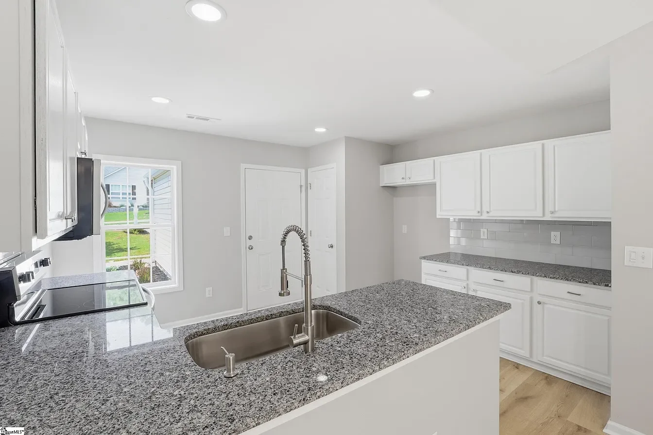 Modern kitchen with granite countertops, white cabinets, stainless steel sink, and a view of a backyard with grass and a house.