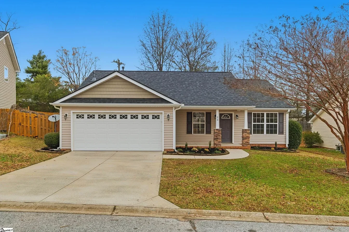 A single-story house with beige siding, a dark gray shingled roof, and a white garage door with decorative windows. The front yard has a lawn, narrow flower beds, and a concrete walkway leading to the front door, which is painted dark purple and flanked by two stone pillars. There are trees with no leaves and a neighboring house in the background.