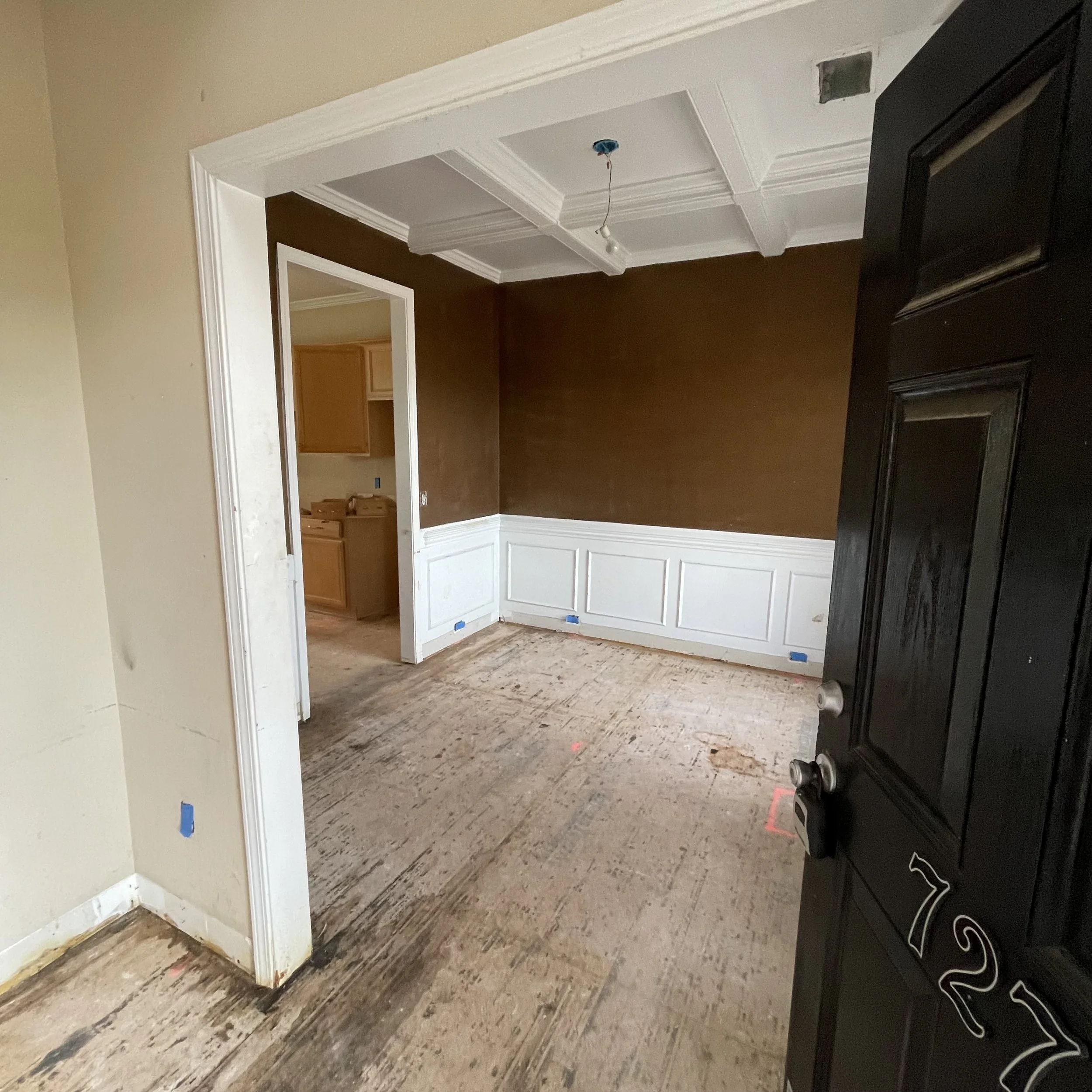 Interior view of a room under renovation, showing unfinished flooring, a doorway, and a kitchen with oak cabinets, with walls painted in various shades, and an open space where a light fixture will be installed.