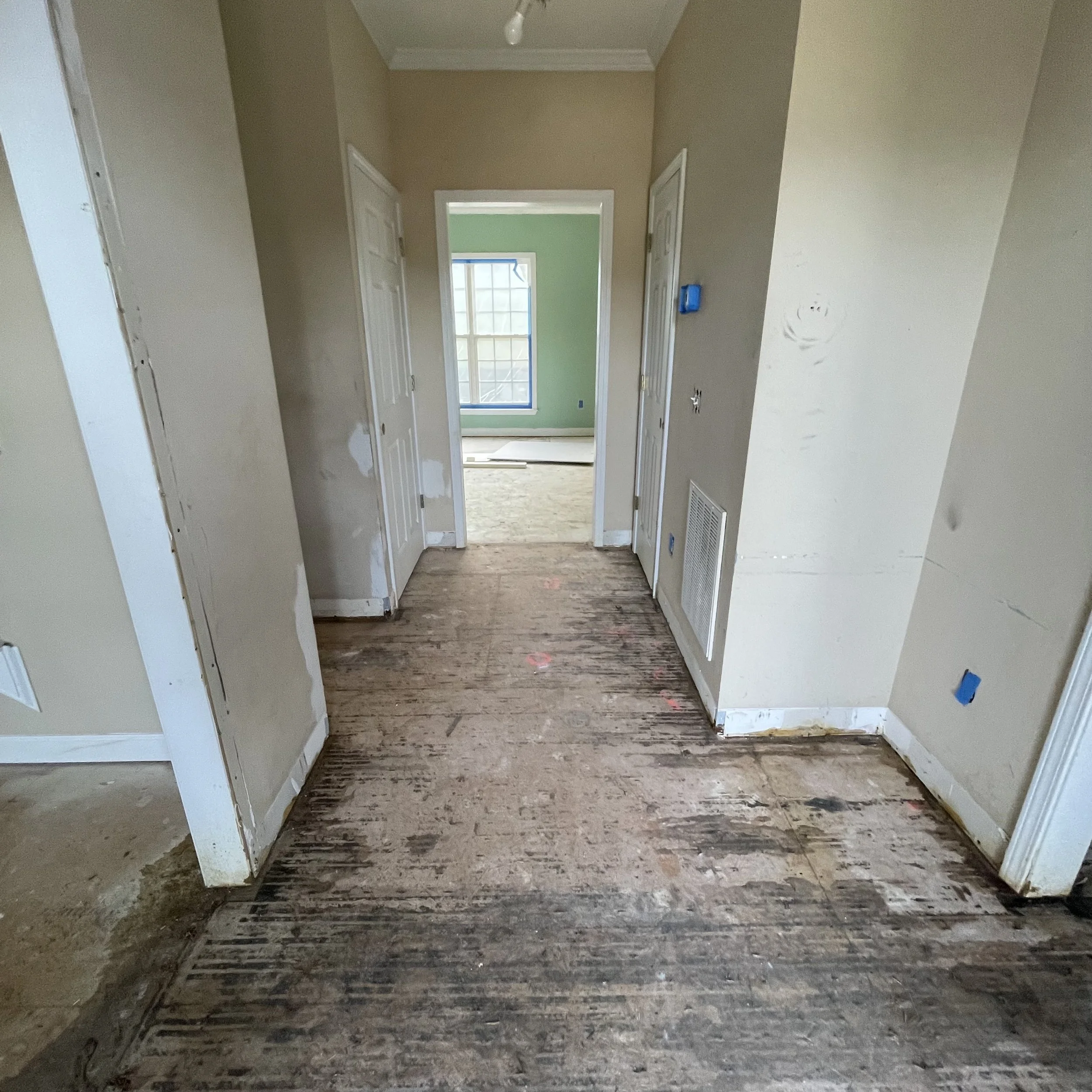 Interior hallway of a house under renovation with exposed wooden floor, white walls, and unpainted door frames leading to rooms with green walls and a window in the background.