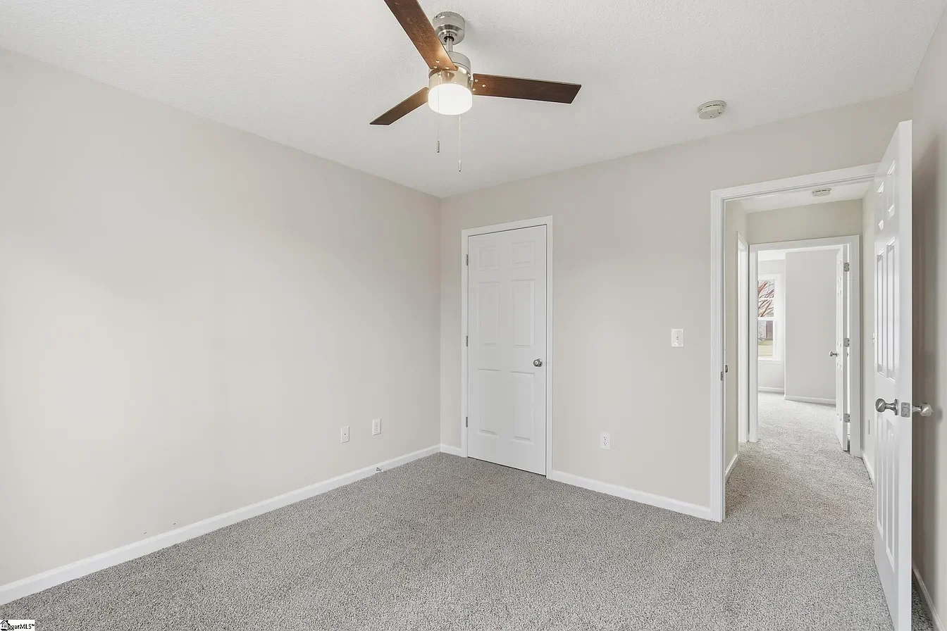 Empty bedroom with beige walls, gray carpet, a ceiling fan with dark blades and white light, and open doors leading to other rooms.