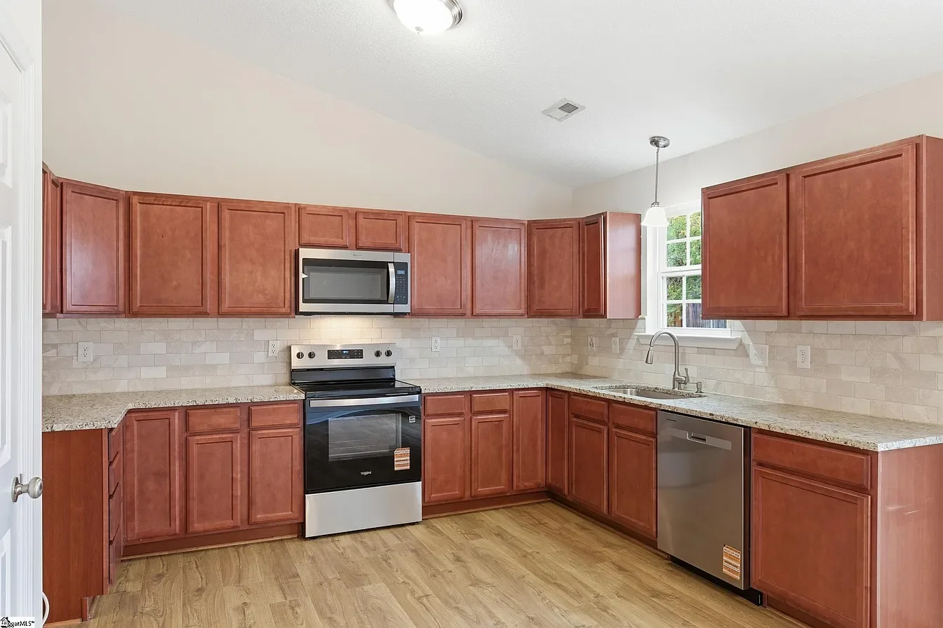 Empty kitchen with wooden cabinets, granite countertops, stainless steel appliances, and a window above the sink.
