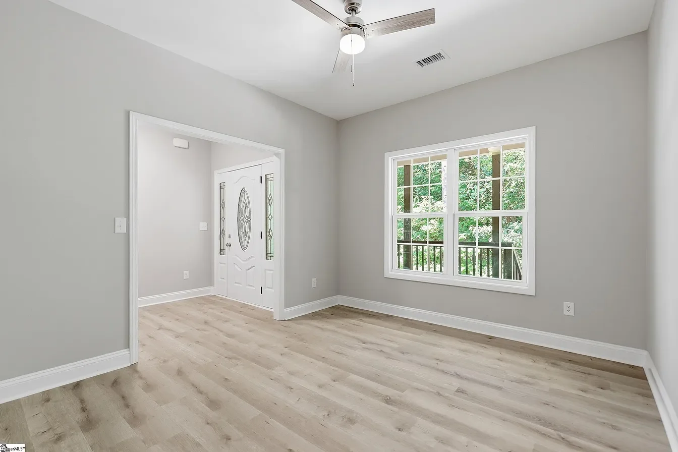Empty room with light wood flooring, gray walls, large window, white front door with decorative glass, and ceiling fan.