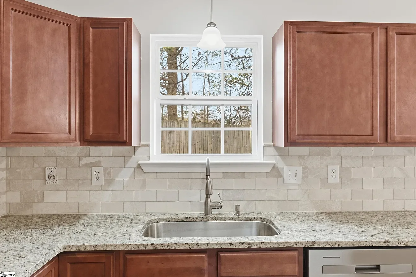 A kitchen window above a granite countertop with a stainless steel sink, brown cabinetry, a white tiled backsplash, and electrical outlets.