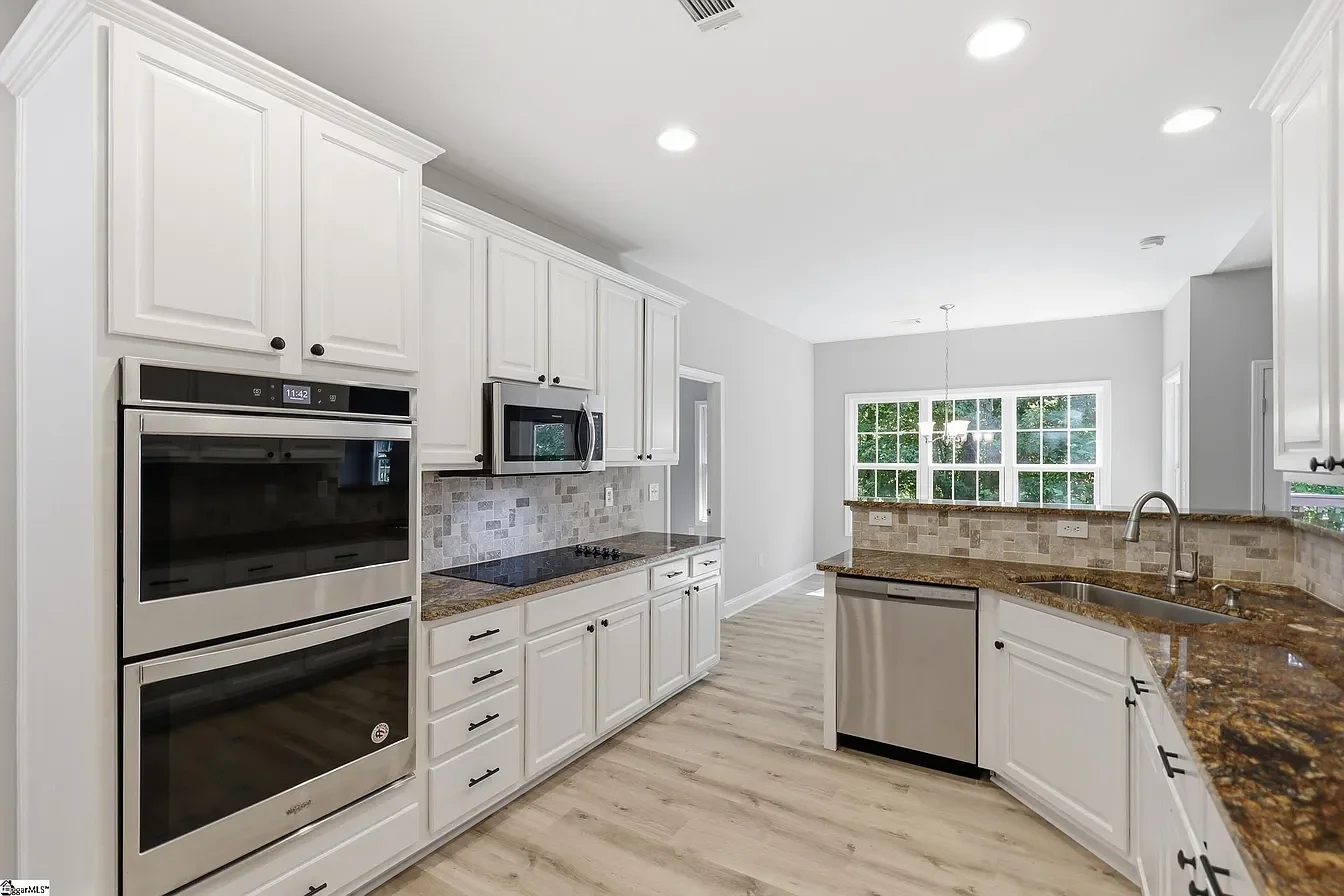 Modern kitchen with white cabinets, granite countertops, stainless steel appliances, and a large window with a dining area in the background.