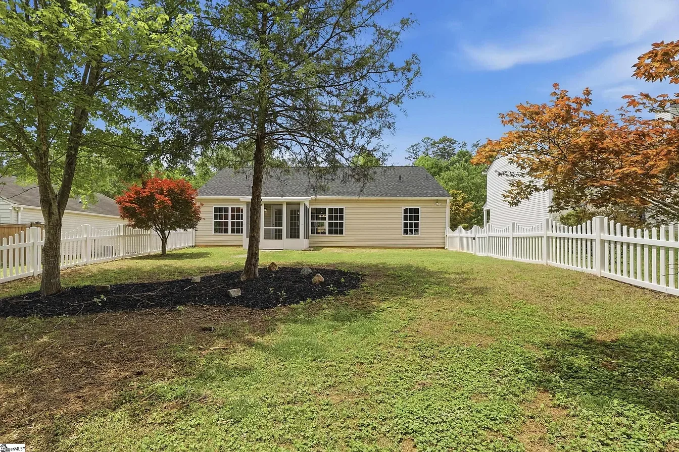 Backyard with green grass, trees, and white fence surrounding the yard. A beige house with multiple windows and a gray roof is visible in the background under a blue sky.