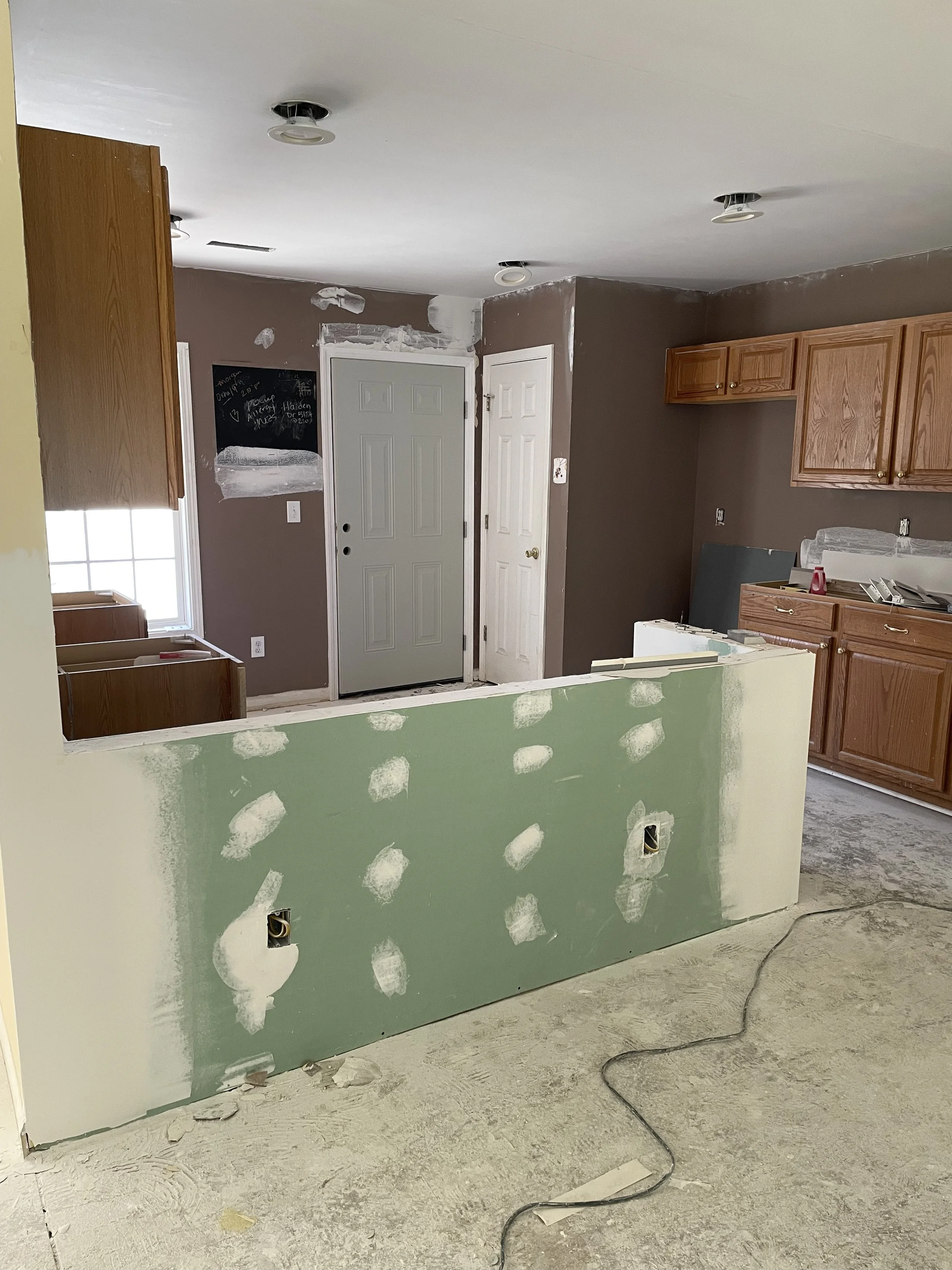 Kitchen under renovation with unfinished drywall, missing cabinet doors, and exposed electrical outlets.