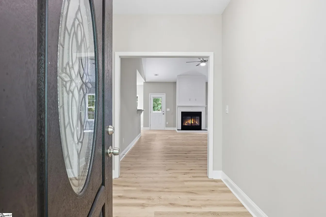 View of a modern living room seen from the slightly open door with decorative glass panel. The room has light gray walls, hardwood floors, a fireplace, a ceiling fan, and a small window.