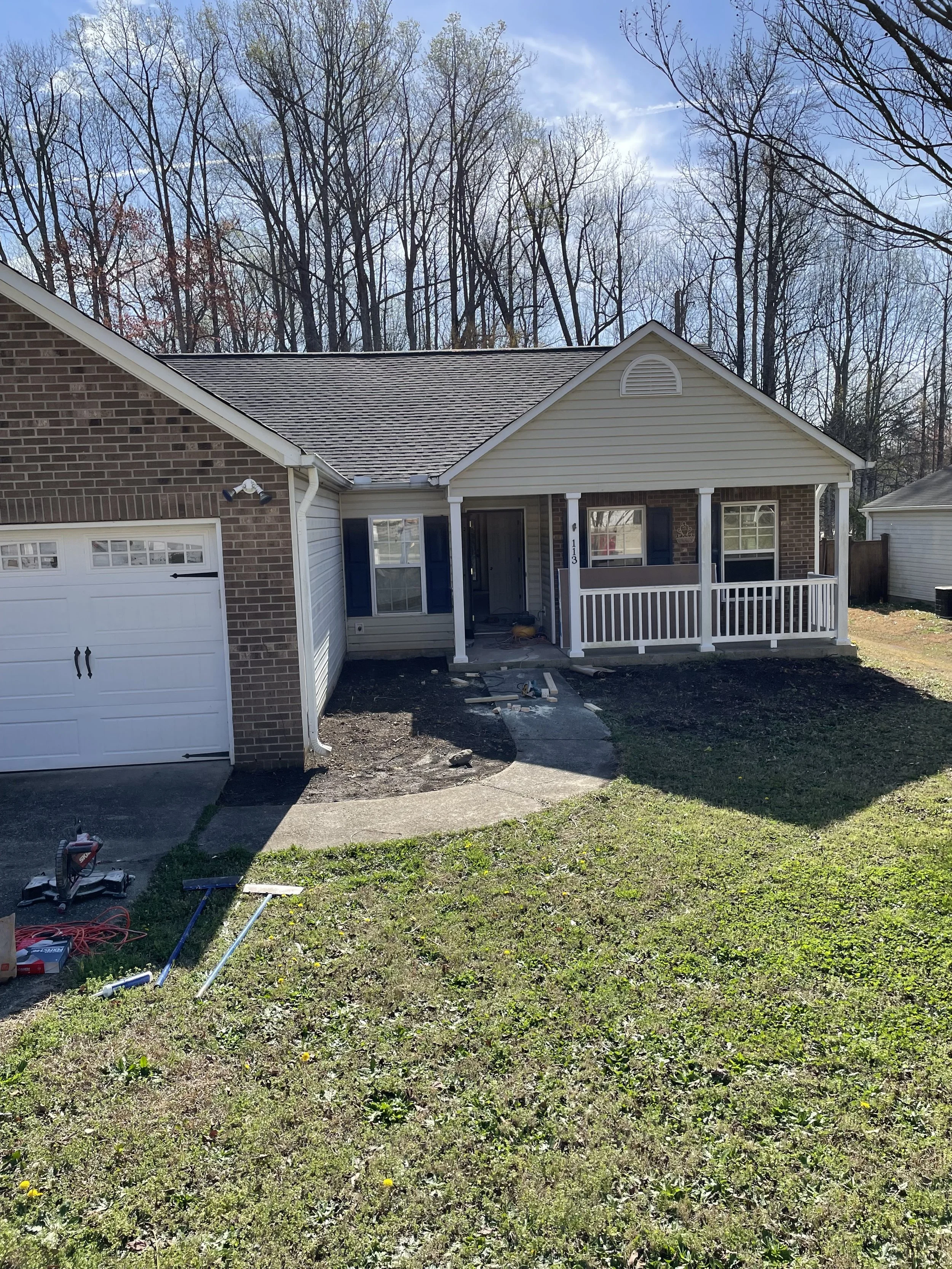 Front yard of a house with a partially renovated porch, tools and construction materials scattered on the grass, a white garage door on the left, and a background of leafless trees under a partly cloudy sky.