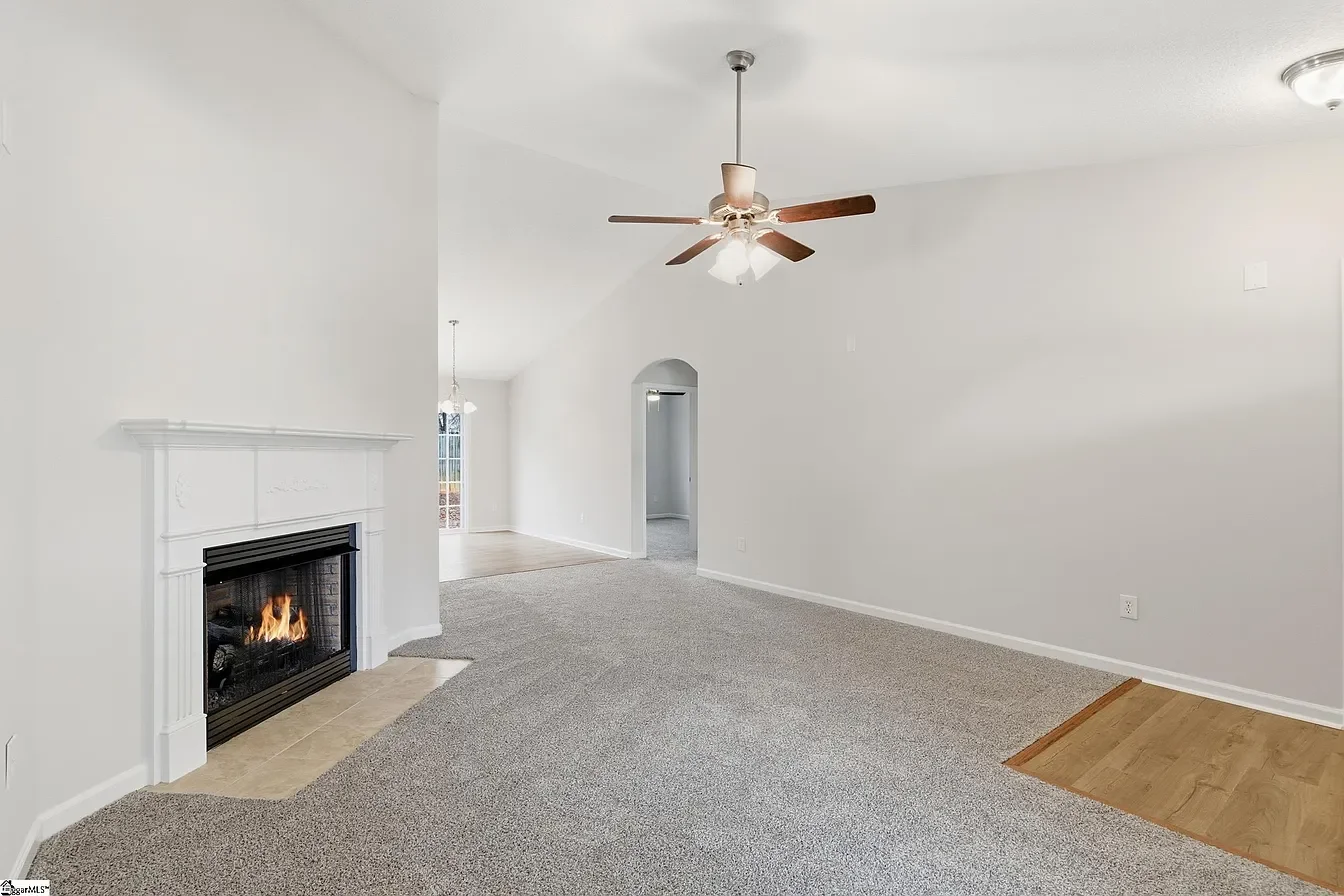 Empty living room with white walls, a gas fireplace, a ceiling fan, and a mix of carpet and hardwood flooring.