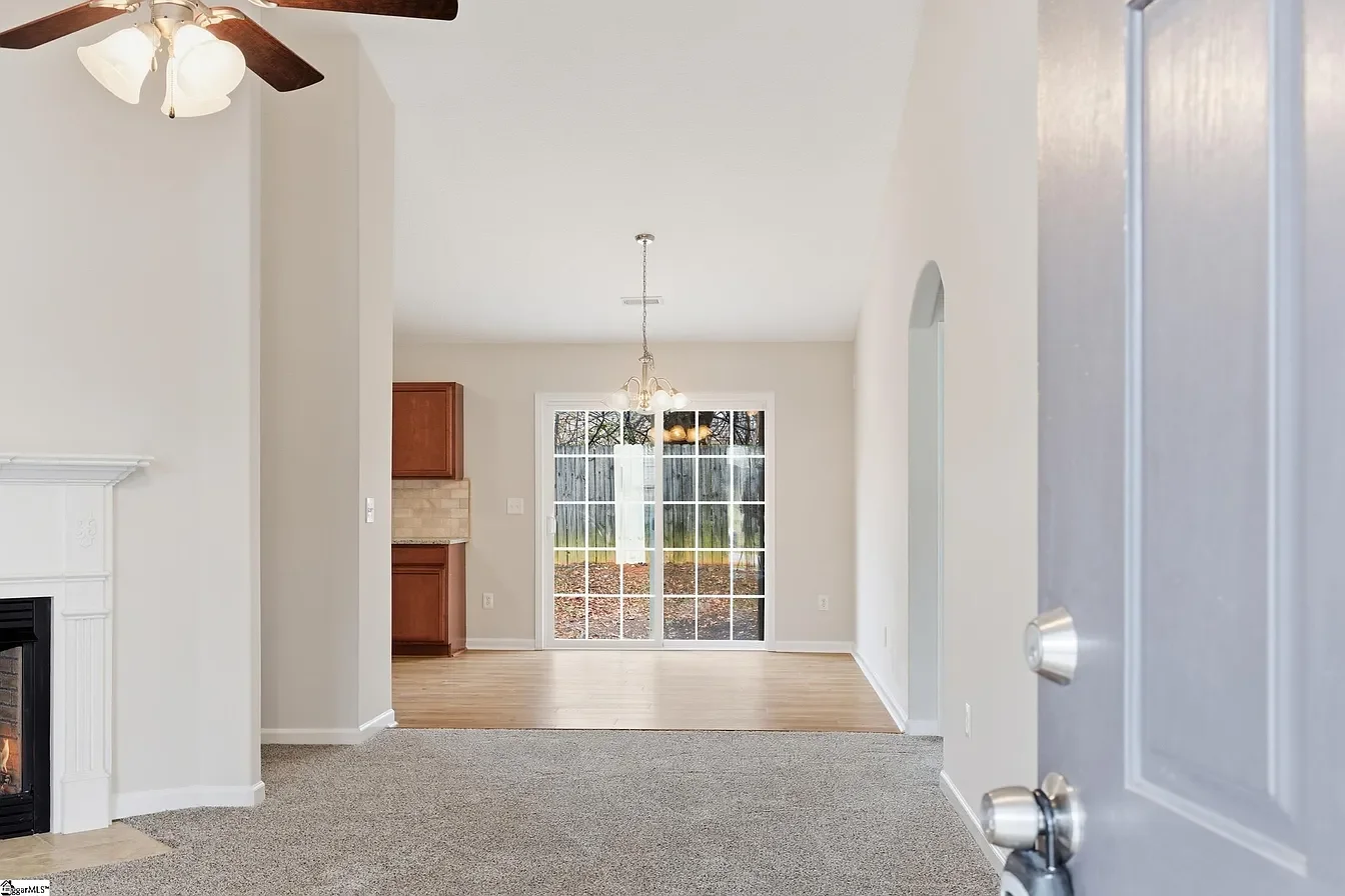 Interior view of a living room and dining area with sliding glass doors leading to an outdoor yard, hardwood and carpet flooring, fireplace, and ceiling fans.