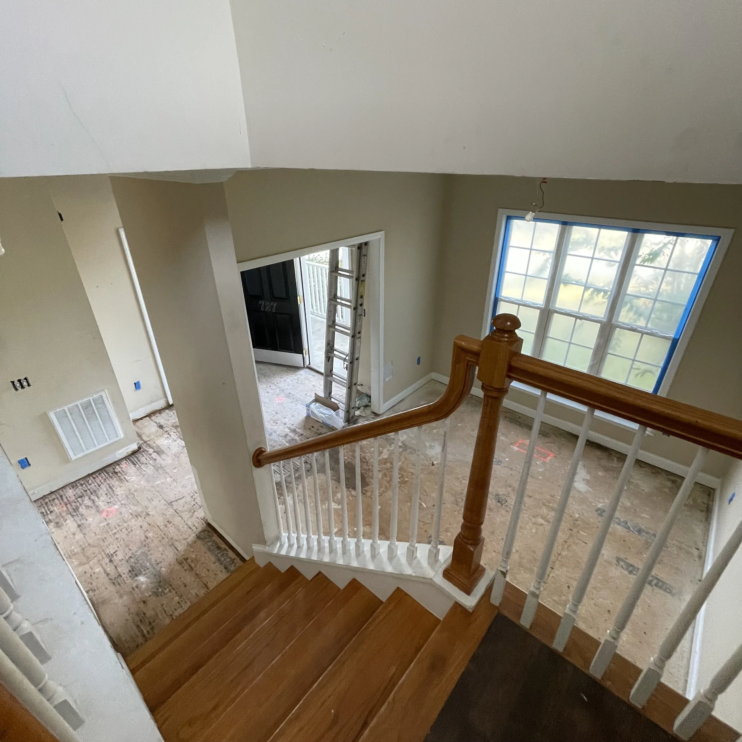A staircase with both wooden and painted white railings, leading to an entryway with a black door and a metal ladder in front of it. The area is under construction or renovation, with visible construction materials and unfinished flooring.