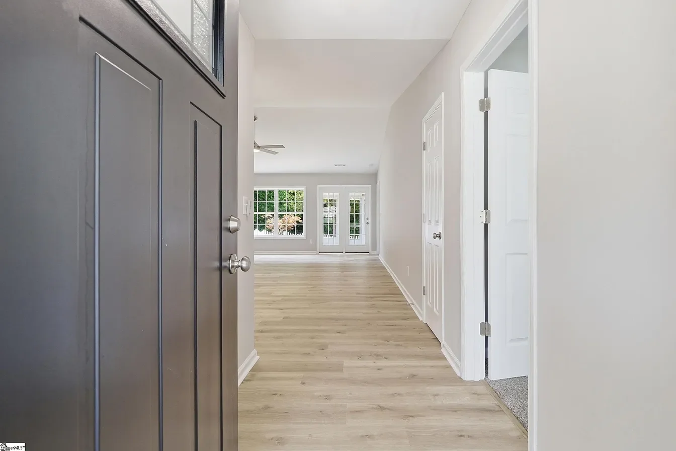 View of an empty, bright living room with large windows and wooden floors, seen from the front door.