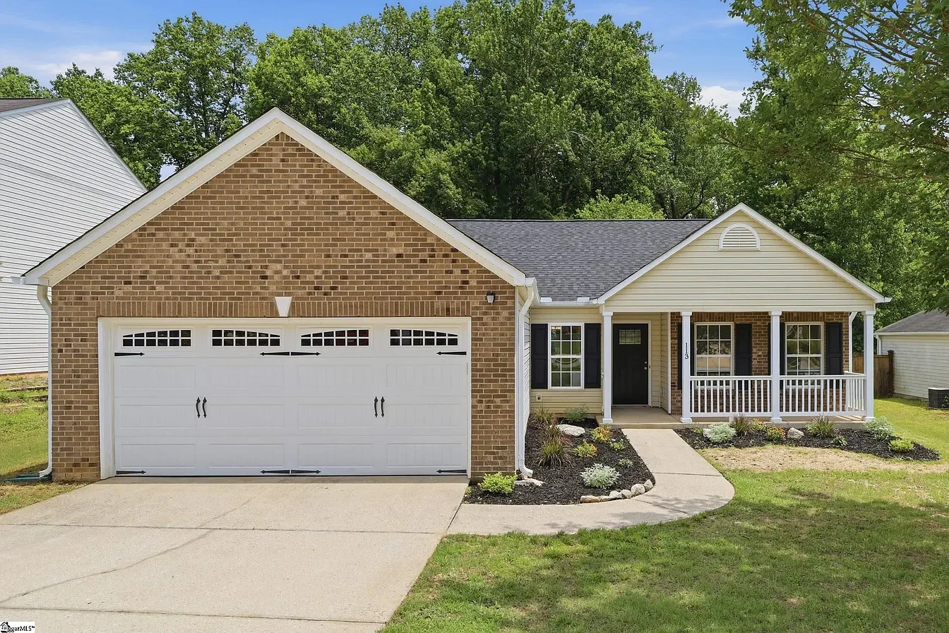 Front exterior of a suburban house featuring a brick garage, a porch with white railings, black shutters, and a landscaped yard with bushes and a walkway.