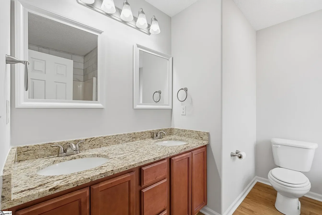 Bathroom with double vanity sink, granite countertop, wooden cabinets, mirror, wall-mounted lights, toilet, and wood flooring.