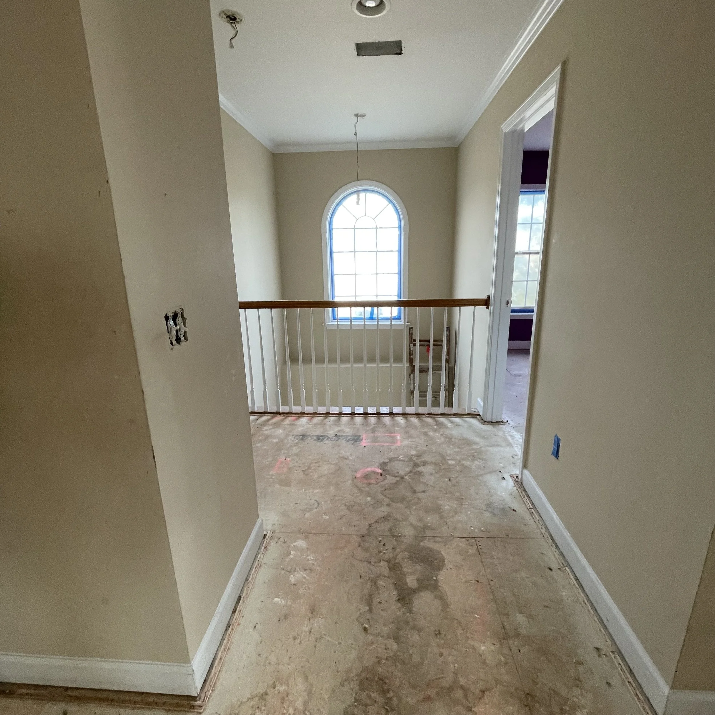 Interior of a house under renovation with an unfinished hallway. The flooring is not yet installed, showing subfloor and debris. There are electrical outlets and wiring visible. The staircase railing is installed, leading to an upper level with a lar