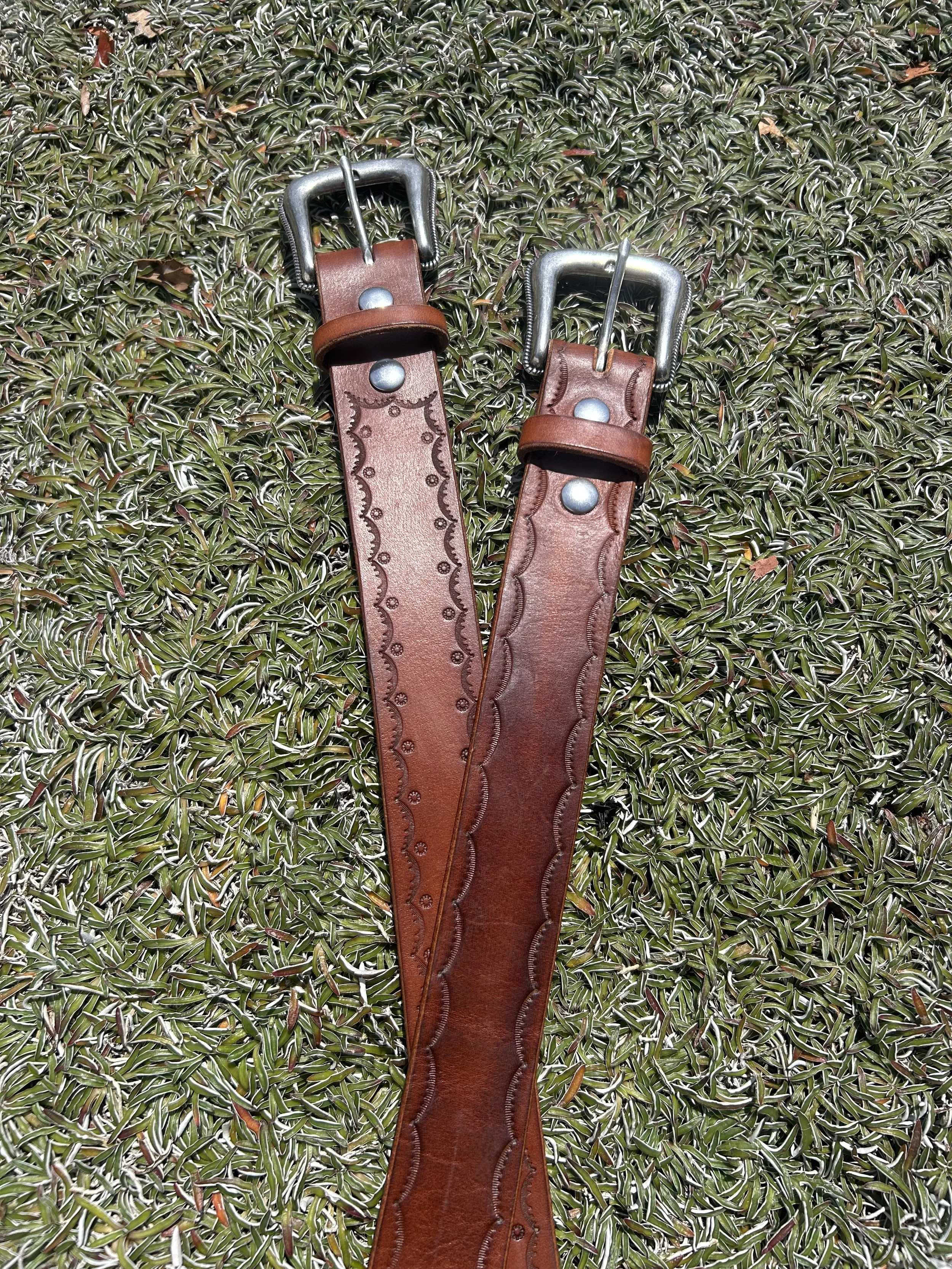 Two brown leather belts with metal buckles lying on green grass.