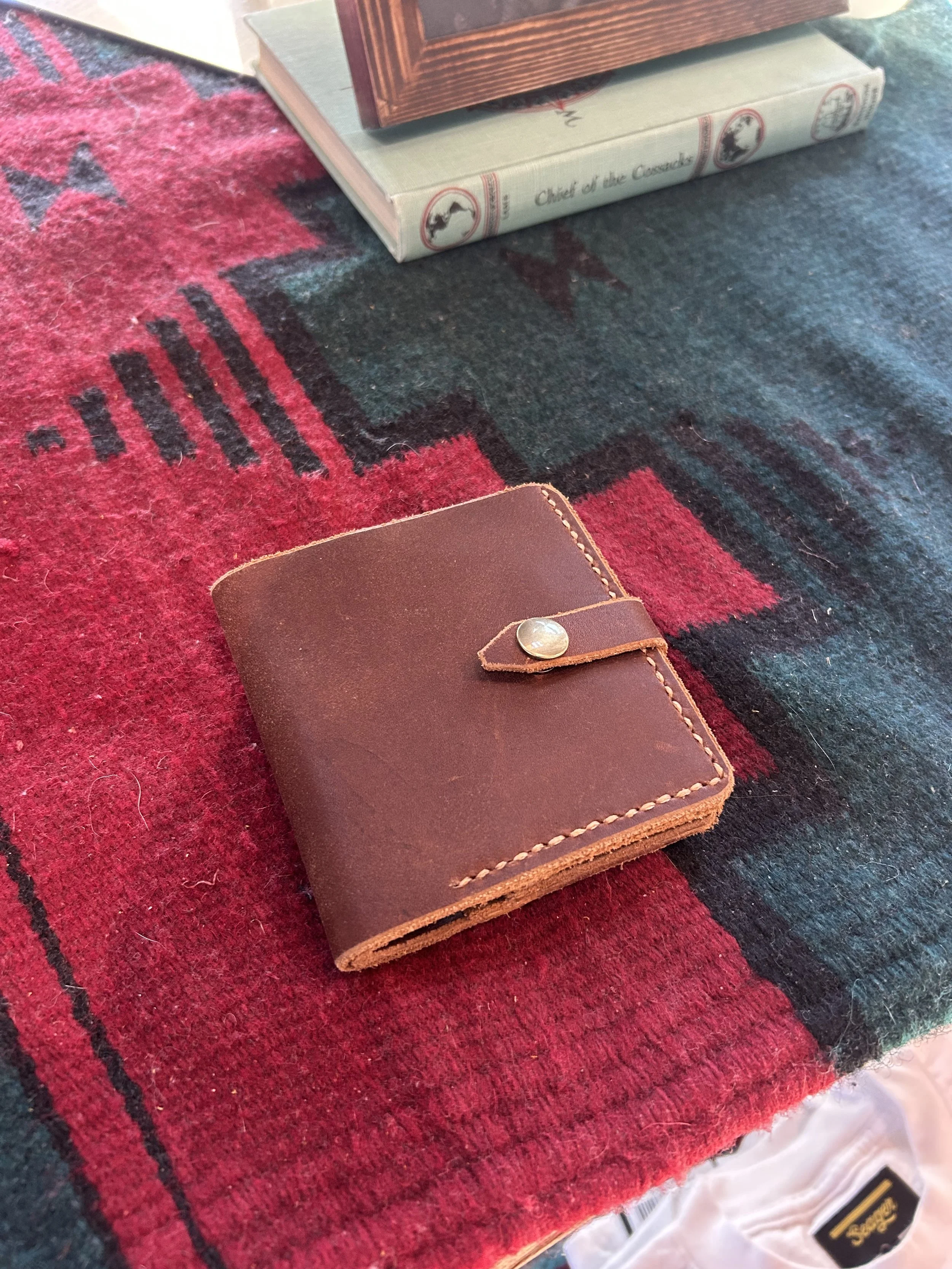 Brown leather wallet with snap button, placed on a red, black, and teal patterned cloth. In the background, there are books and a wooden frame.