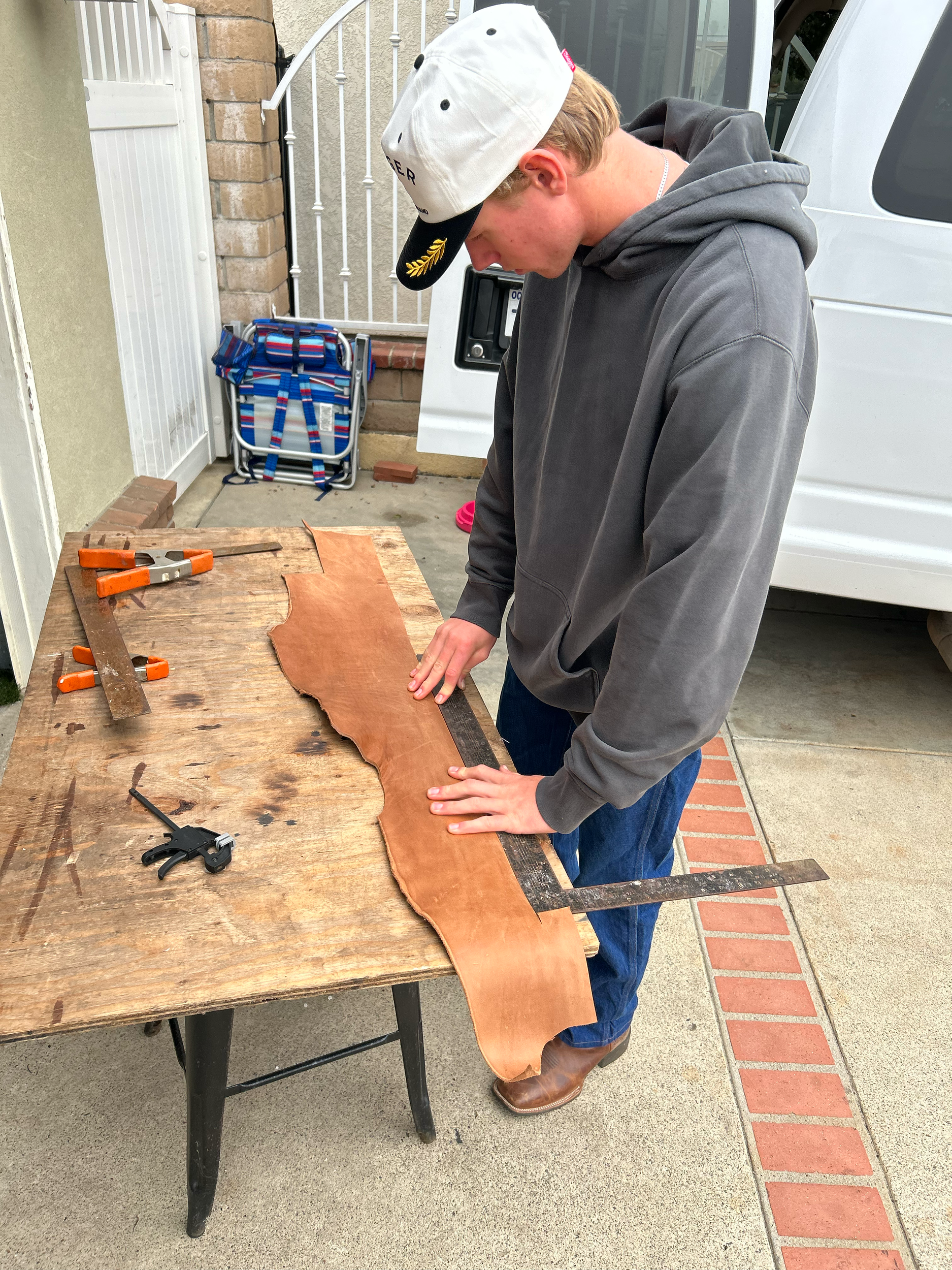 A young man working on a piece of leather on a wooden table, using clamps and a ruler, outdoors near a white van.