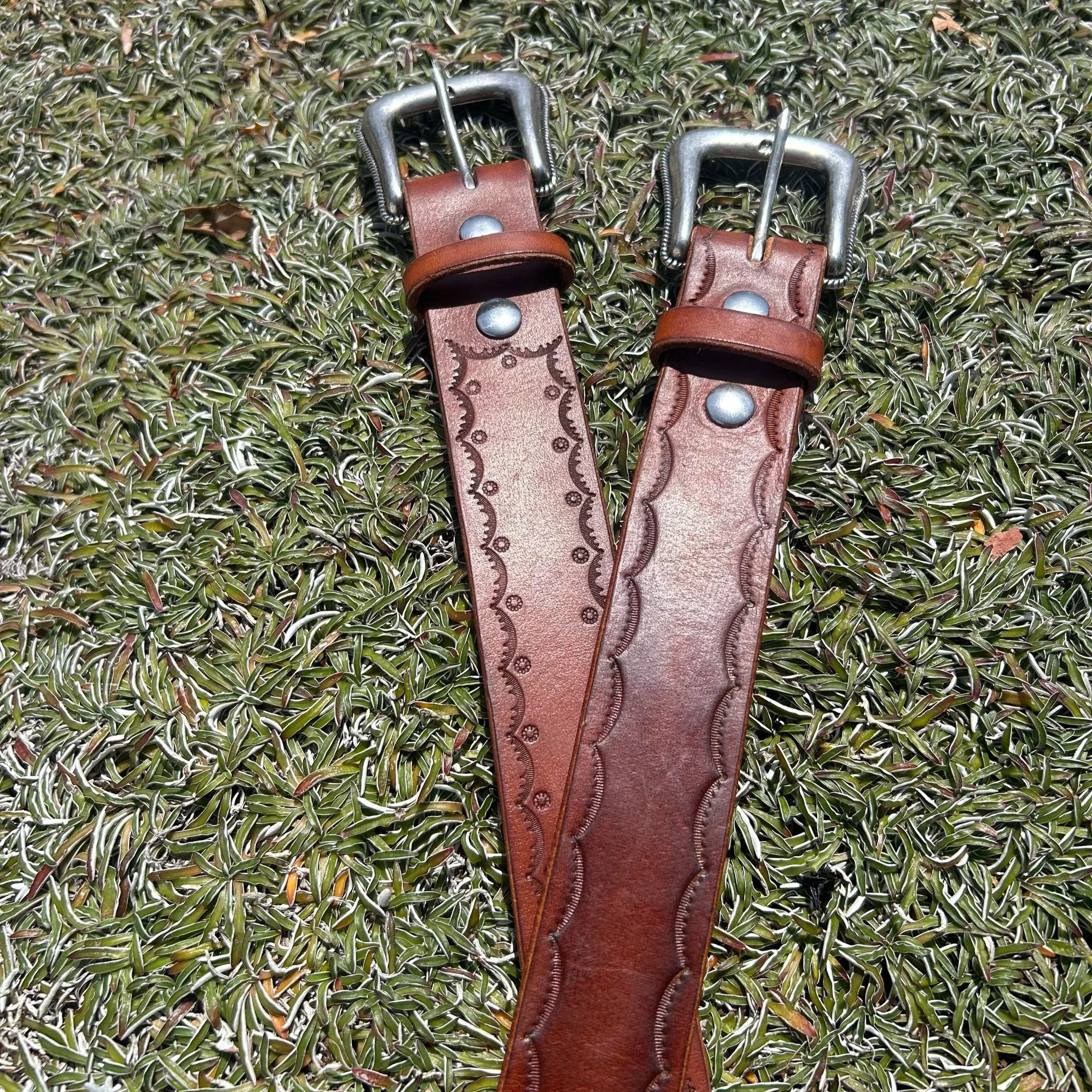 Two brown leather belts with decorative stitching and metal buckles lying on green grass.