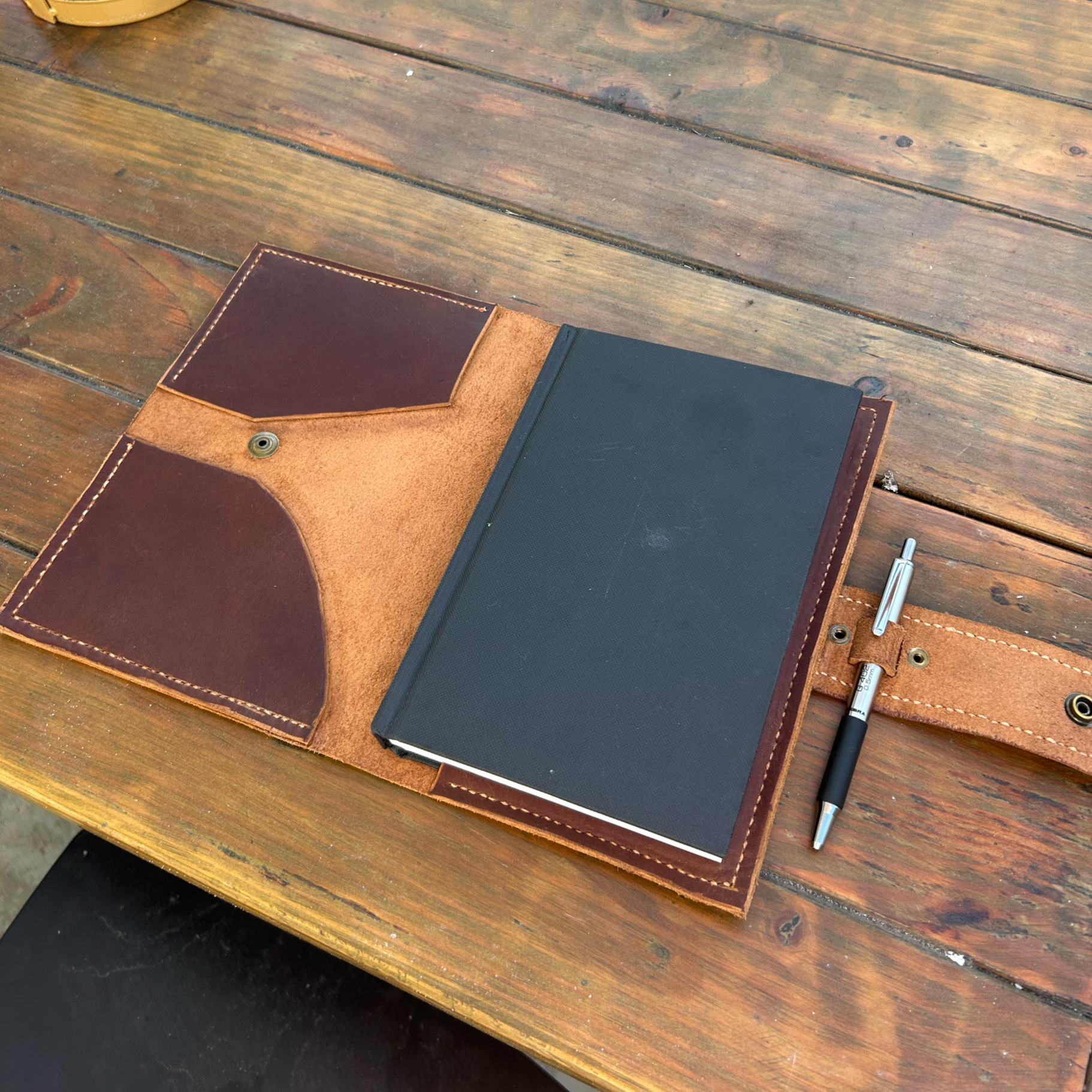 An open leather wallet with a black notebook inside, resting on a wooden table, with a silver and black pen nearby.