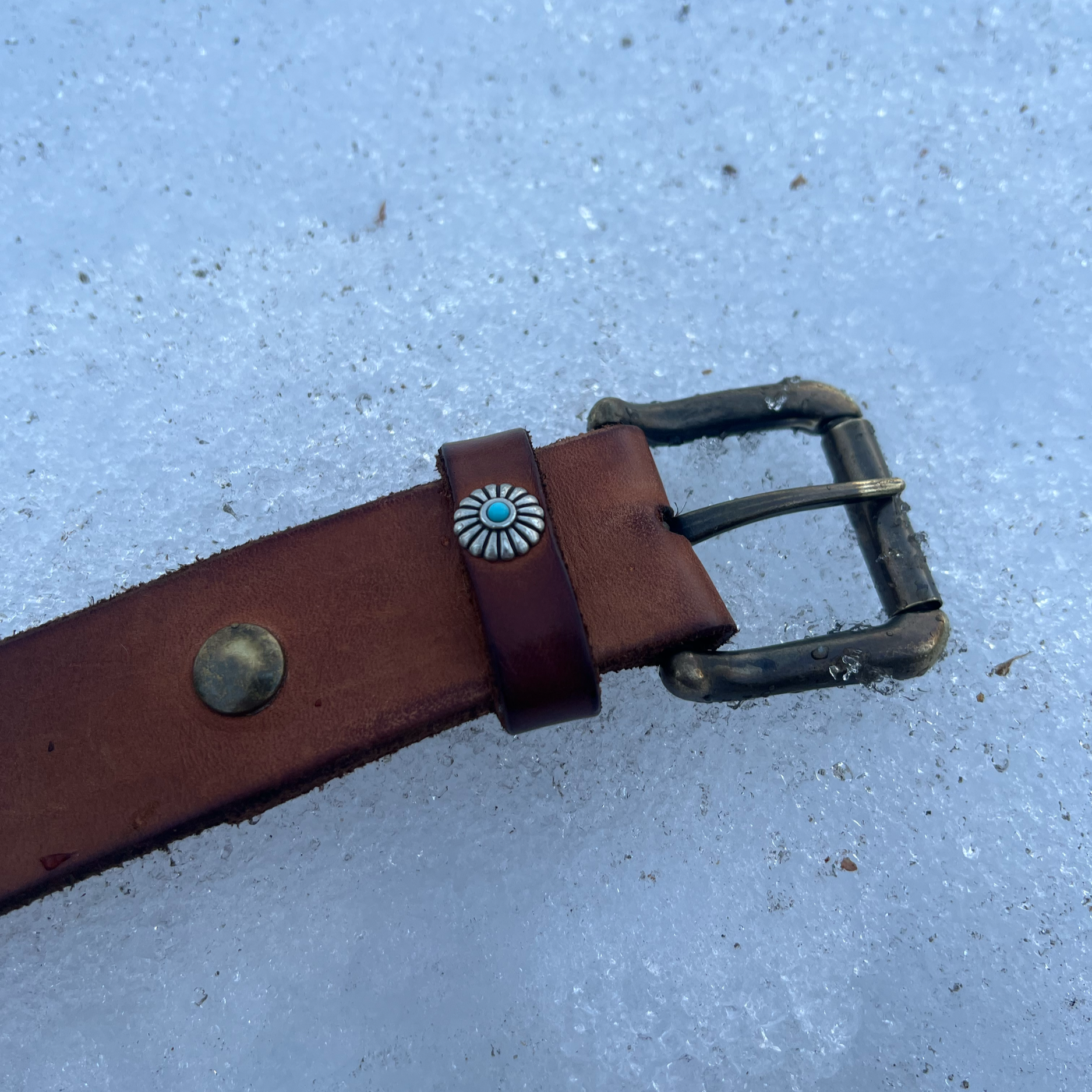 Close-up of a brown leather belt with a decorative silver and turquoise concho, lying on snow with a metal buckle.