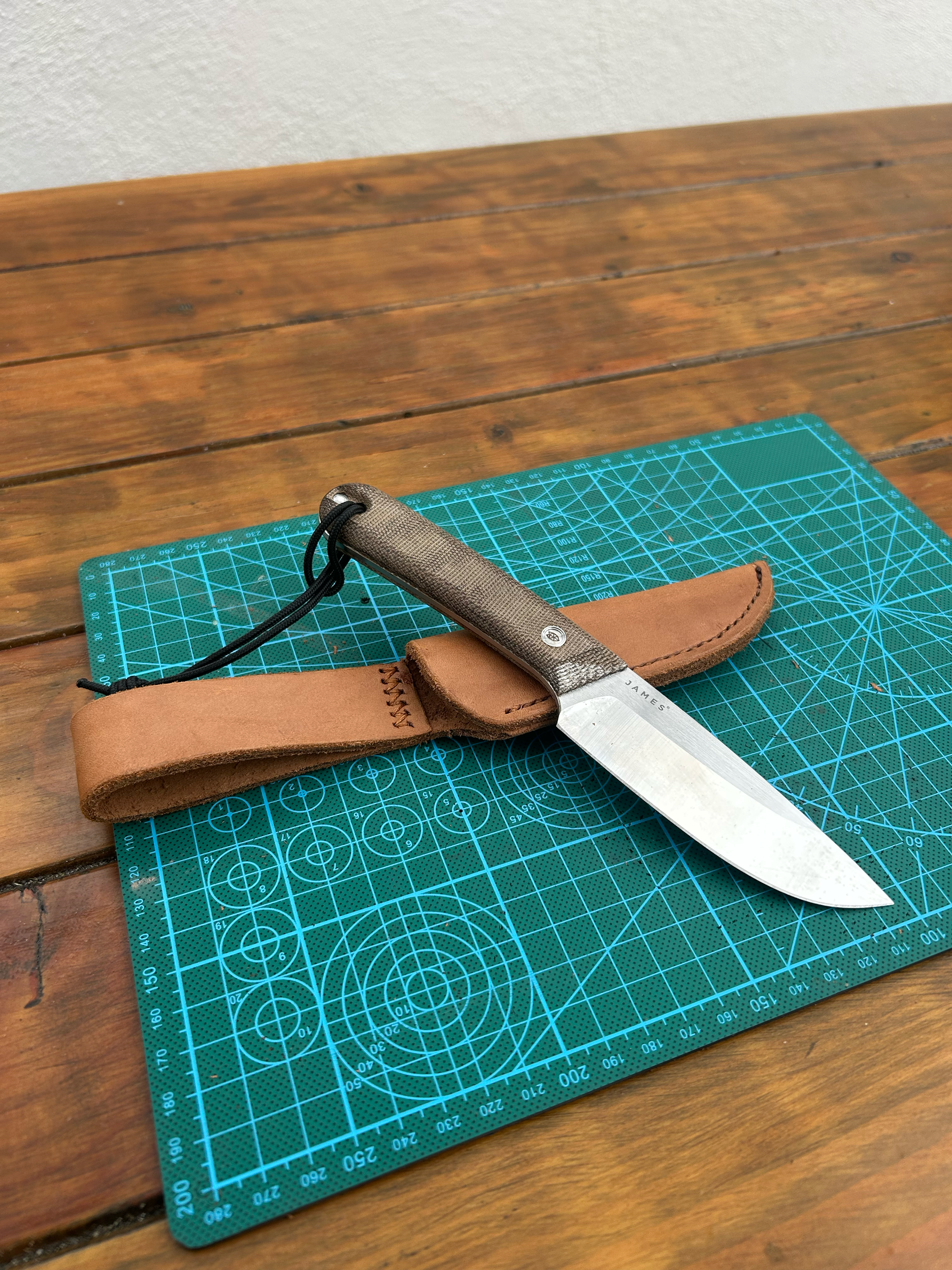 A fixed-blade knife with a black lanyard, resting on a leather sheath, on top of a green cutting mat on a wooden table.