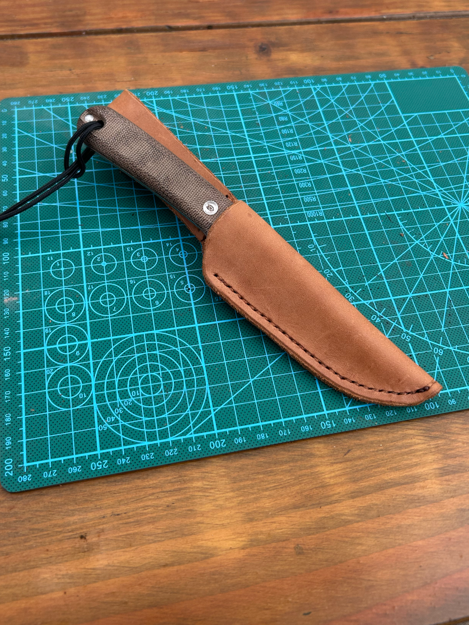 A fixed blade knife with a brown leather sheath and a fabric handle, lying on a blue cutting mat on a wooden surface.