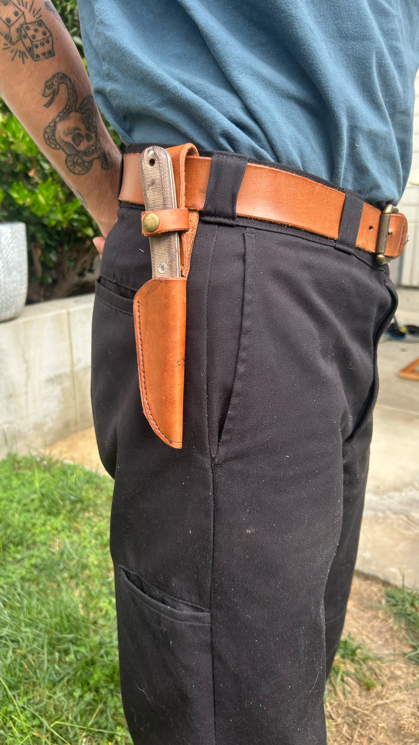 Close-up of a person wearing black pants, a brown leather belt, and a knife with a textured handle in a leather sheath attached to the belt, speaking to a tattooed arm, outdoors with grass and a concrete block wall in the background.