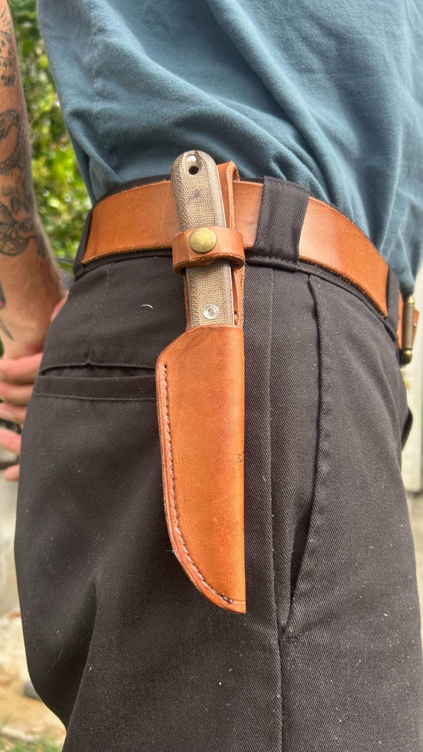 A person wearing black pants and a brown leather belt, with a fixed-blade knife in a brown leather sheath attached to the belt.