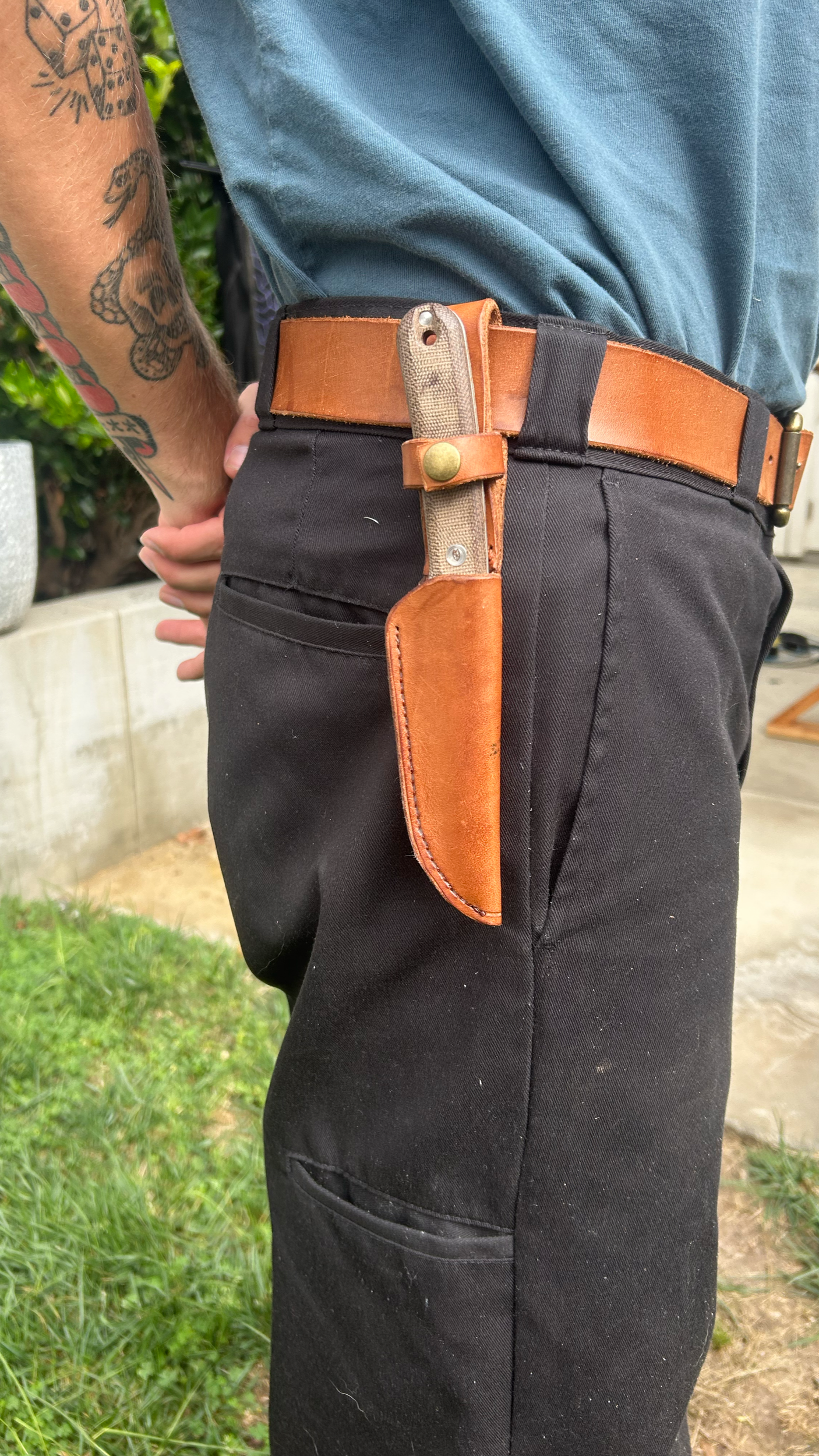 A person with tattoos on their arm wearing a blue shirt and black pants, with a brown leather belt holding a fixed-blade knife in a leather sheath attached to the side.