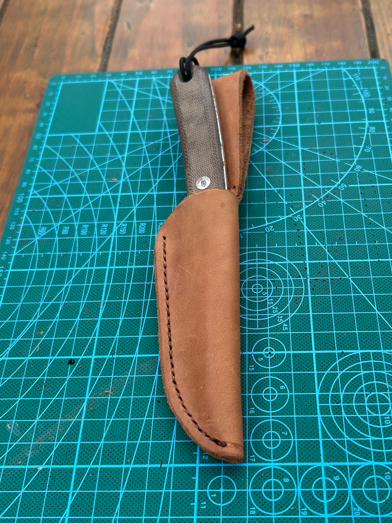 A folding pocket knife with a textured dark handle and a silver bolt, partially sheathed in a brown leather case, resting on a green cutting mat with measurement grid.