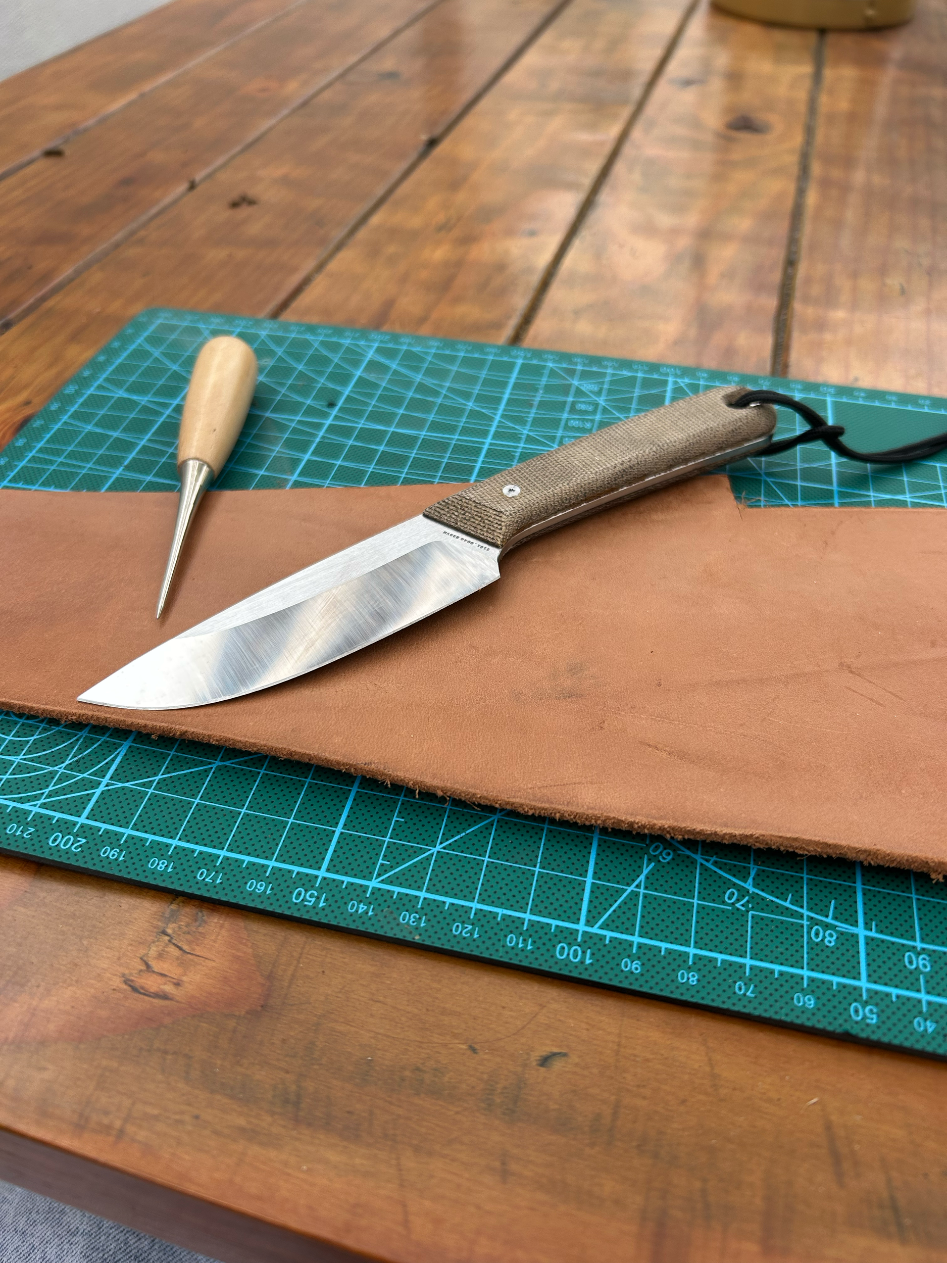 A craft knife and a leather punch tool resting on a piece of tan leather, with a green cutting mat and a wooden table in the background.