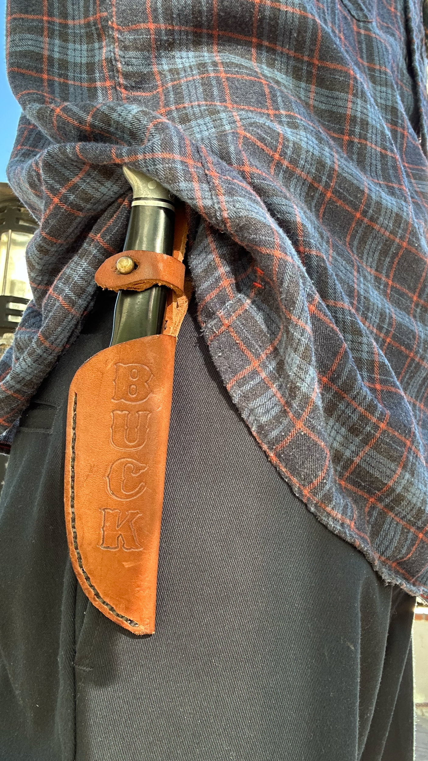 A person wearing a dark plaid shirt and black pants has a pocket knife in a brown leather sheath attached to their belt.