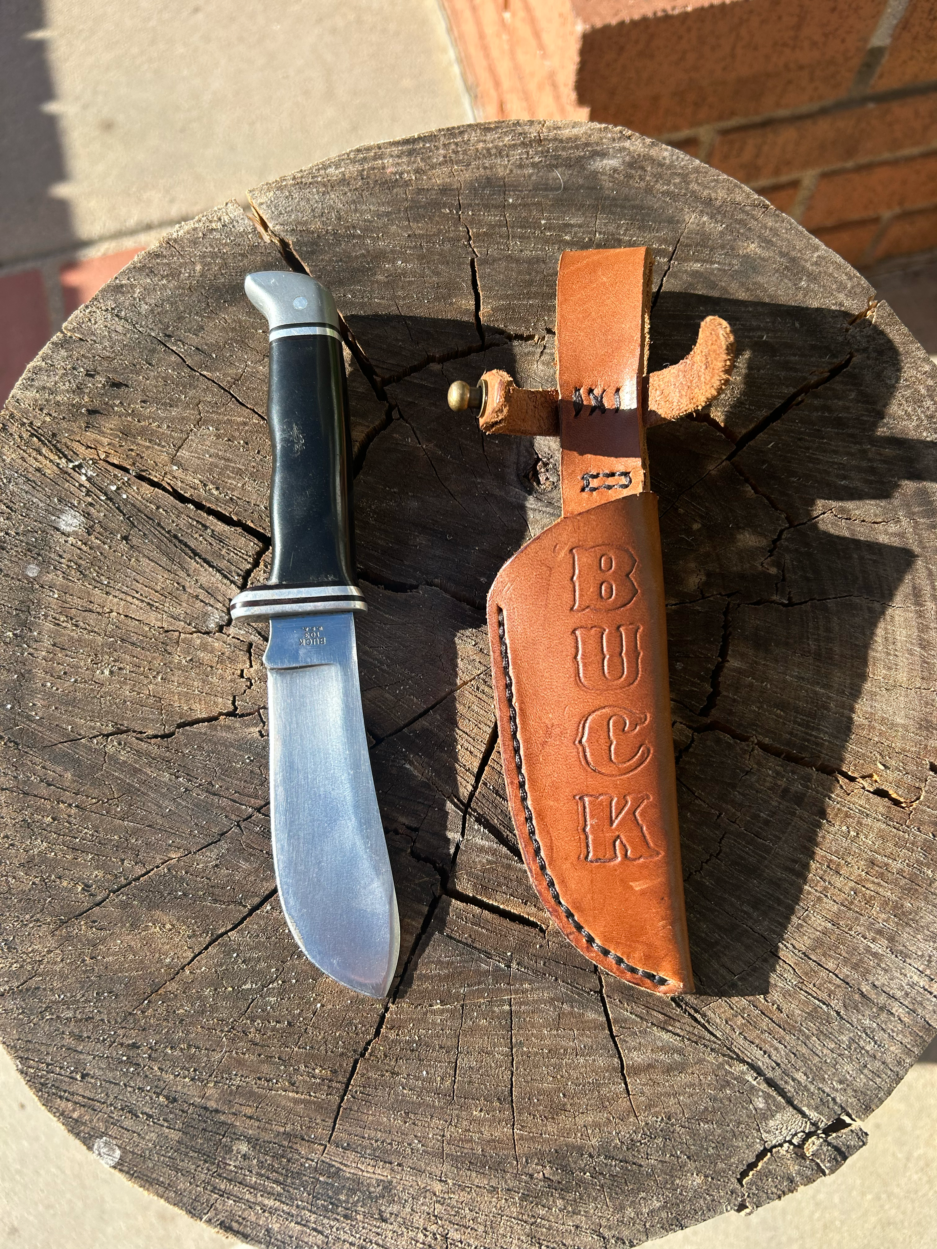 A folding pocket knife with a black handle and metal blade placed on a weathered wooden surface, with a brown leather sheath embossed with the words 'BUCK' beside it.