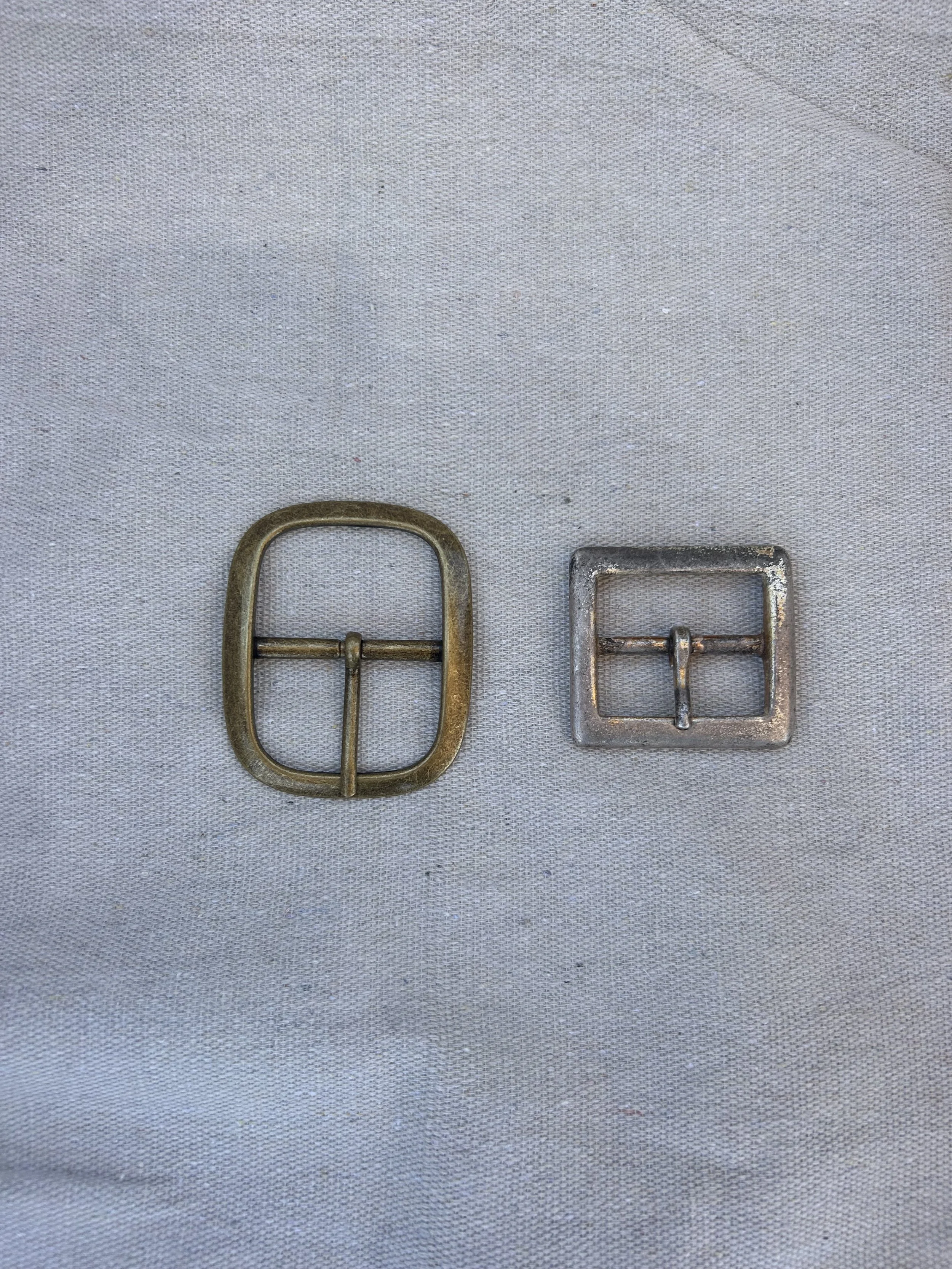 Two metal belt buckles, one brass-colored and oval-shaped, the other silver-colored and square-shaped, placed on a gray fabric background.