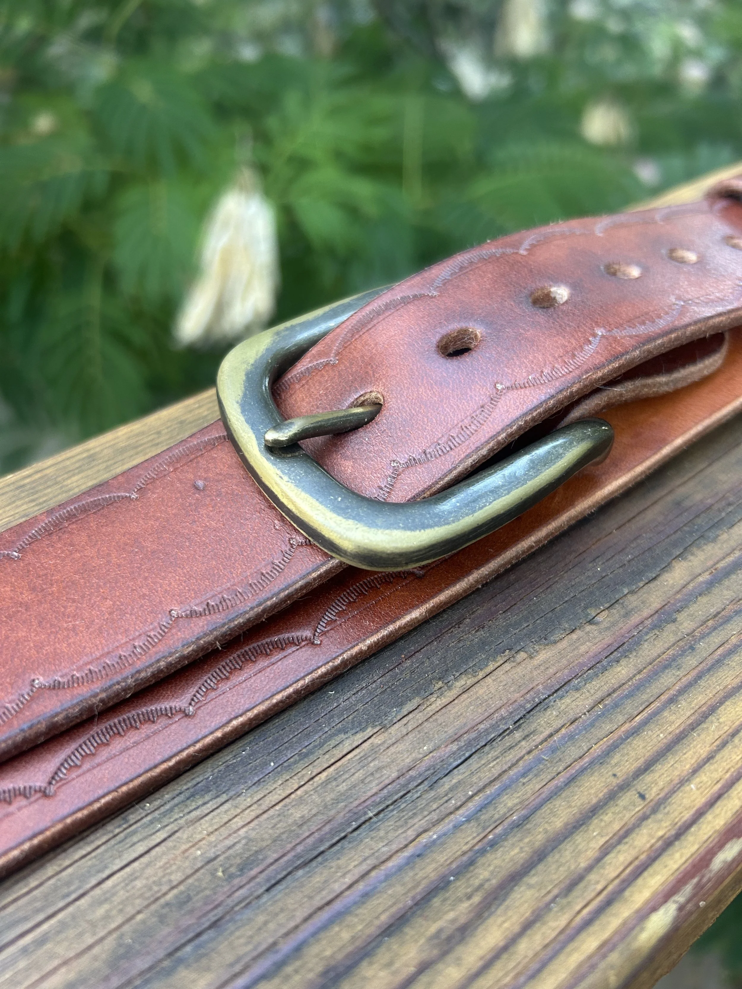 Close-up of a brown leather belt with a brass buckle lying on a wooden surface, with greenery in the background.