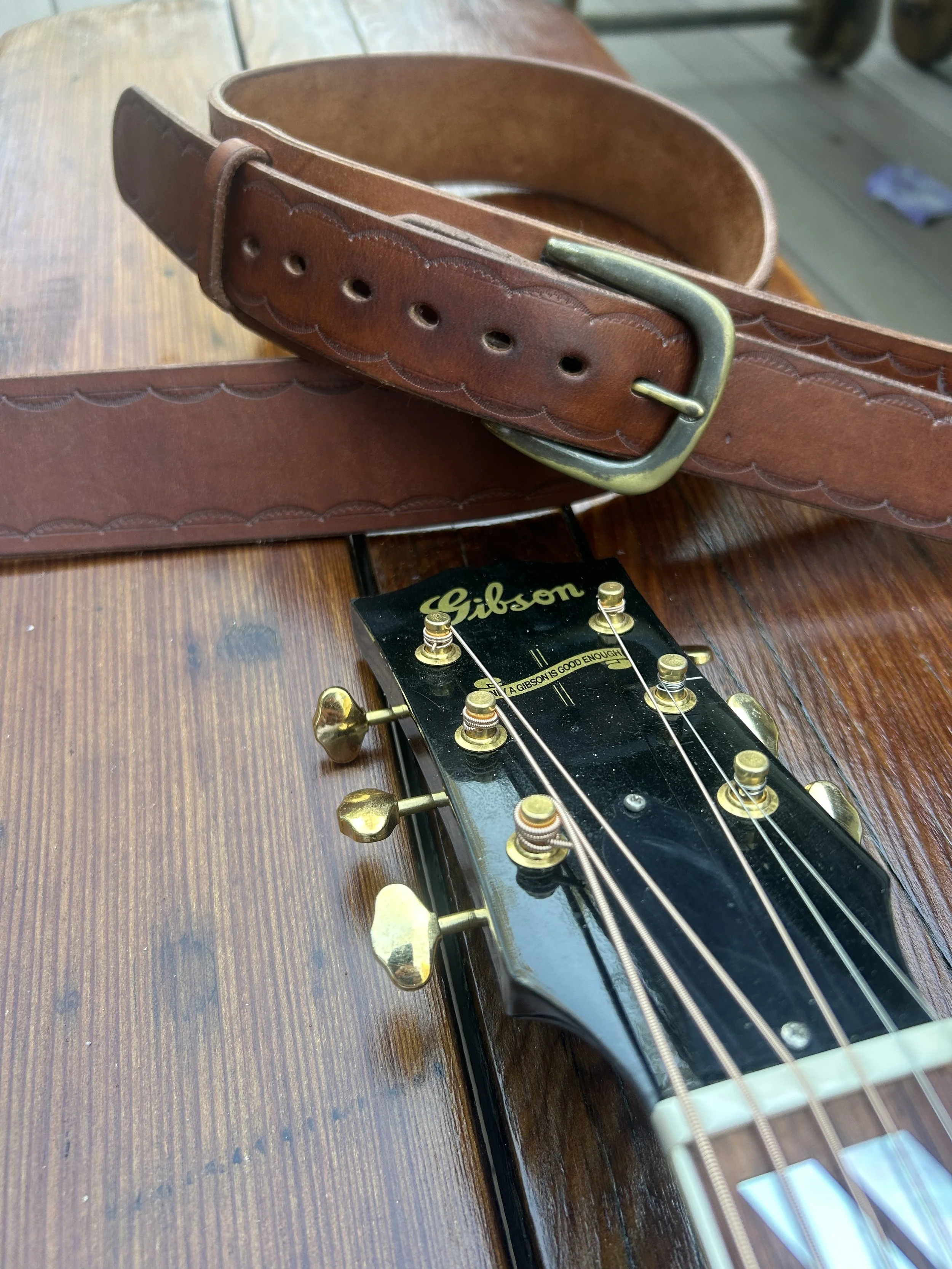 Close-up of a Gibson electric guitar headstock with tuning pegs, lying on a wooden surface, with a worn brown leather belt in the background.
