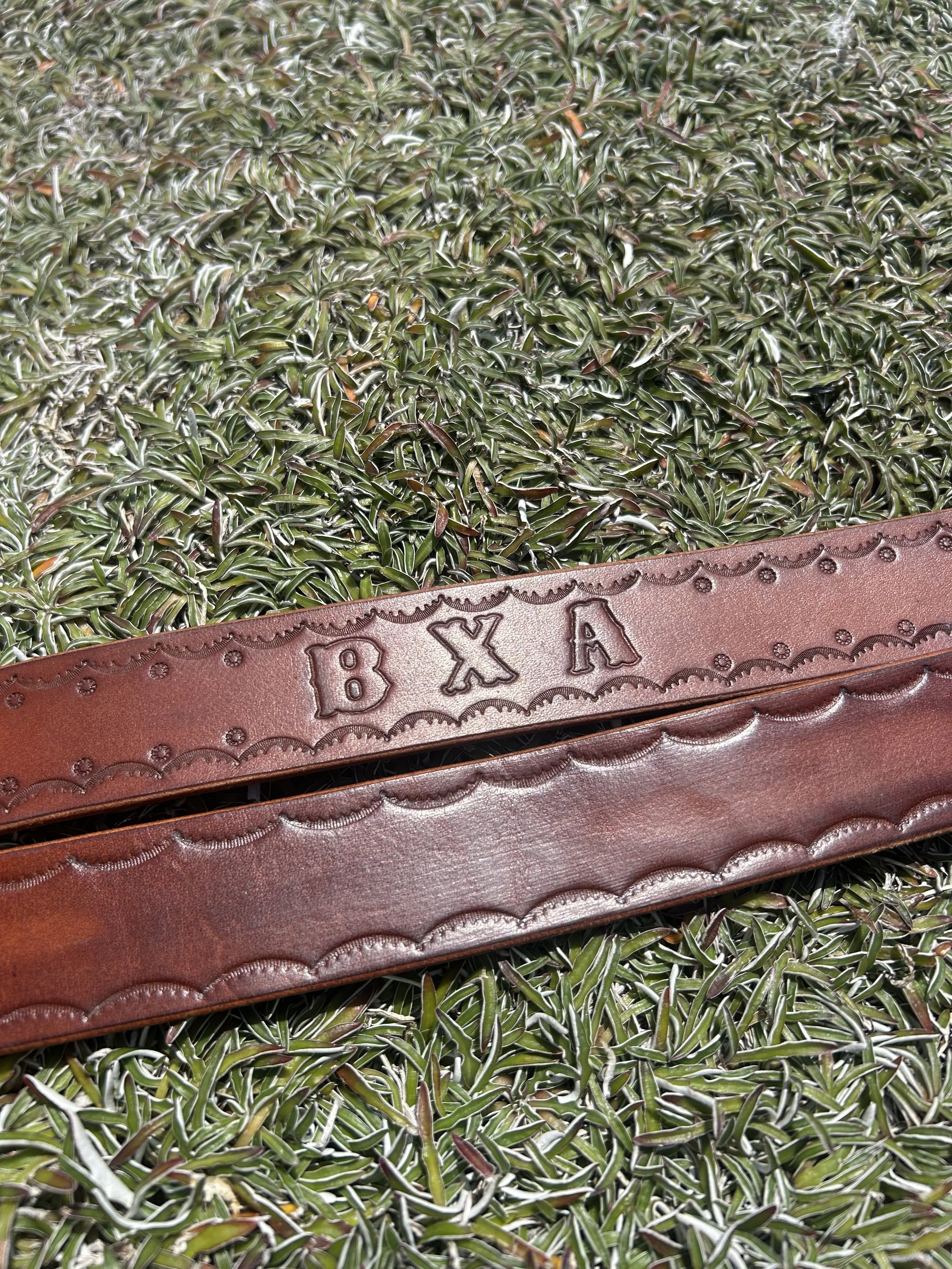 A brown leather belt with the embossed letters 'B X A' lies on a bed of green and silver foliage.