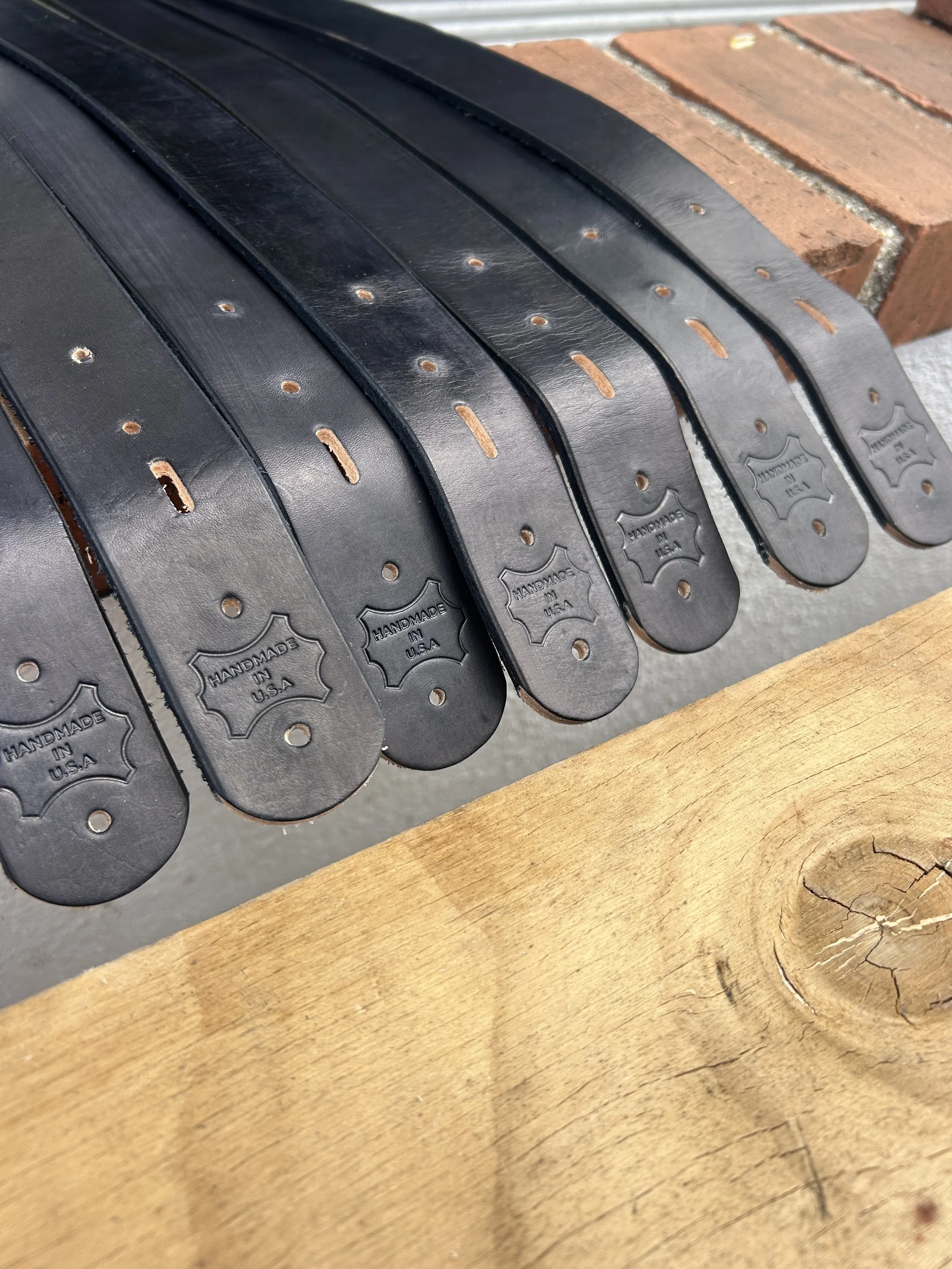 Several black leather belts with stamped "Handmade in U.S.A." logos, laying on a wooden surface.