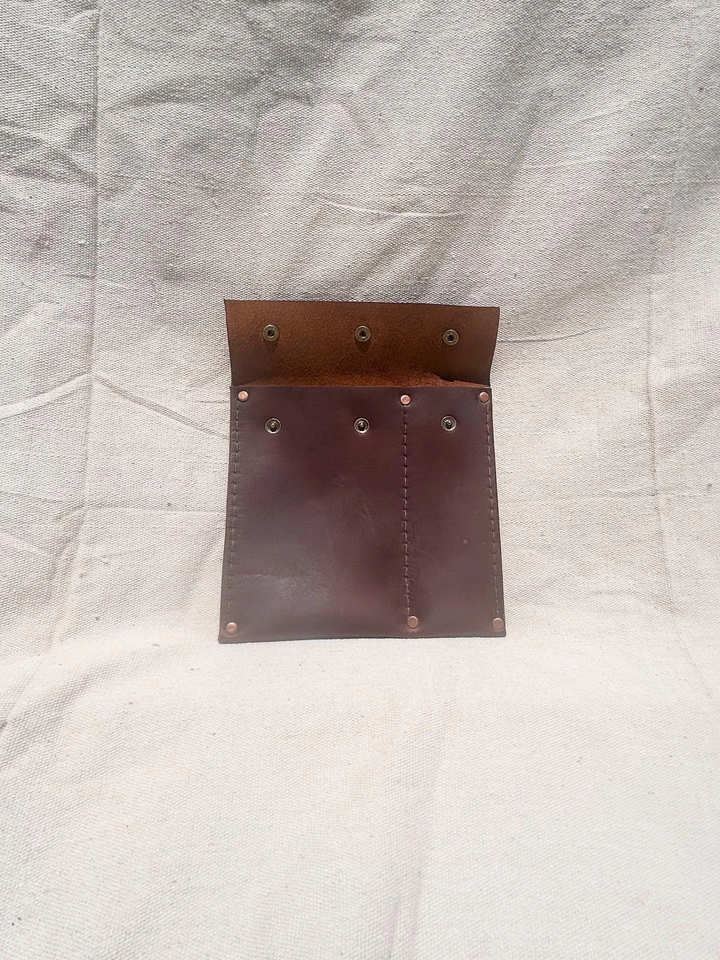 A brown leather wallet with multiple rivets and compartments, open and placed on a white fabric surface.