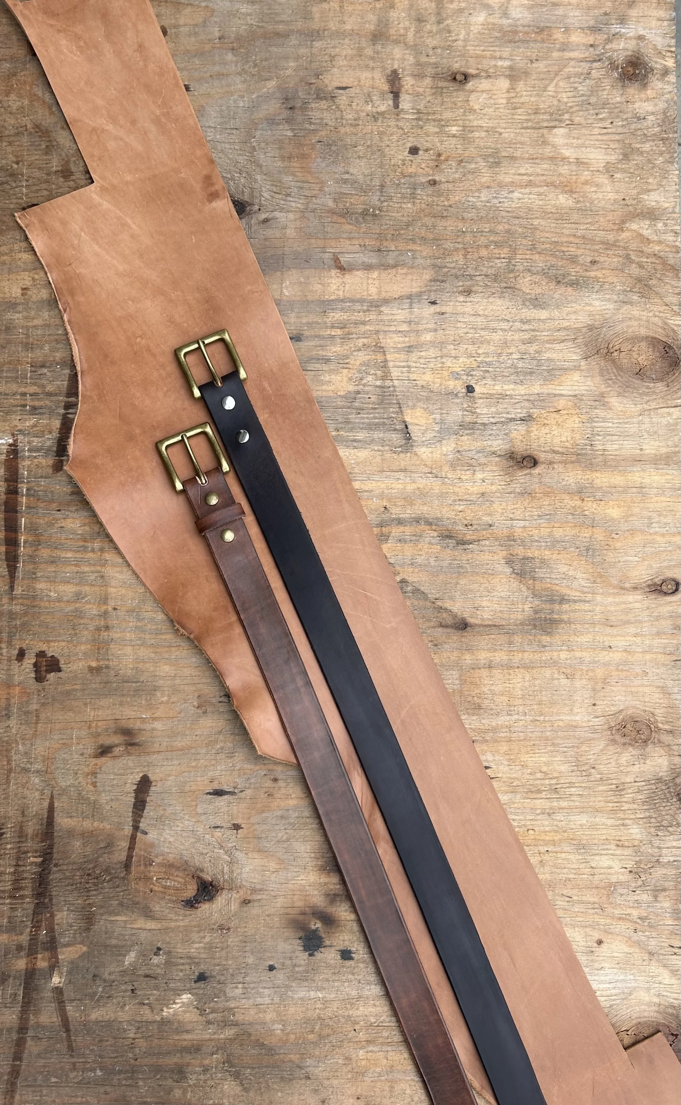 Two leather belts, one dark brown and one black, with brass buckles, placed on a piece of light brown leather on a wooden surface.