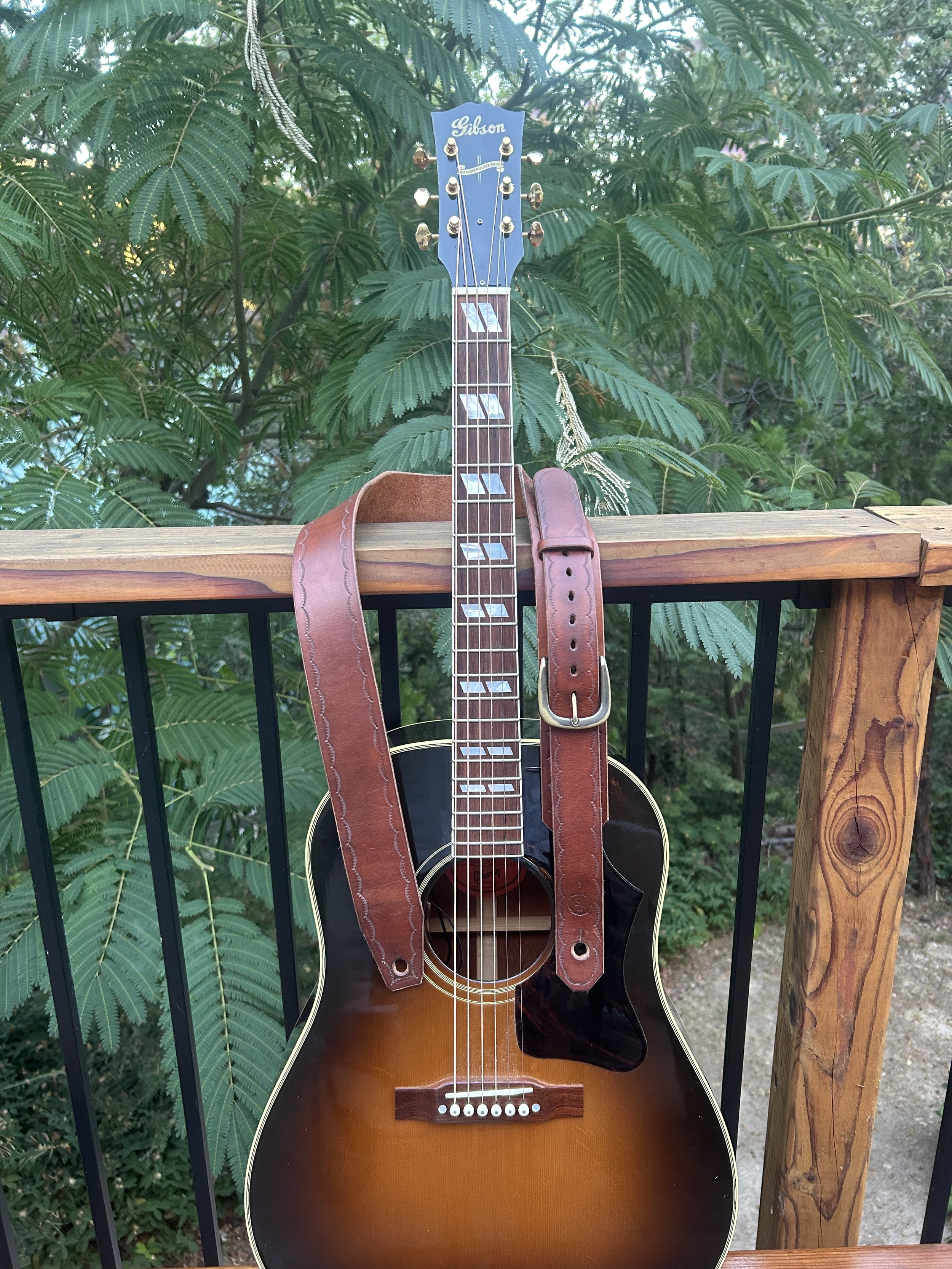 An acoustic Gibson guitar with a brown strap hanging on a railing, with green leafy foliage in the background.