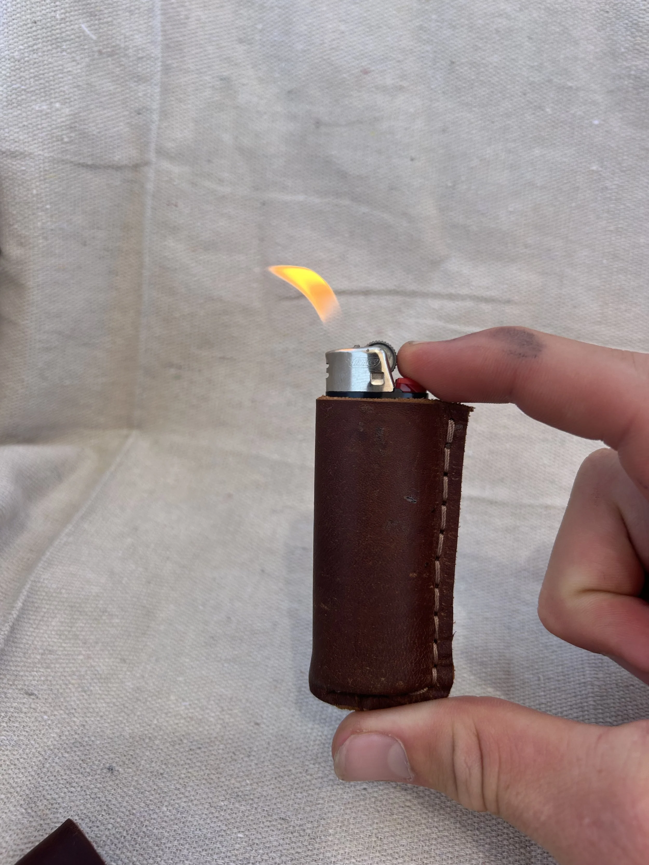 A hand holding a lighter with a small flame, with a brown leather sleeve around the lighter, against a beige fabric background.