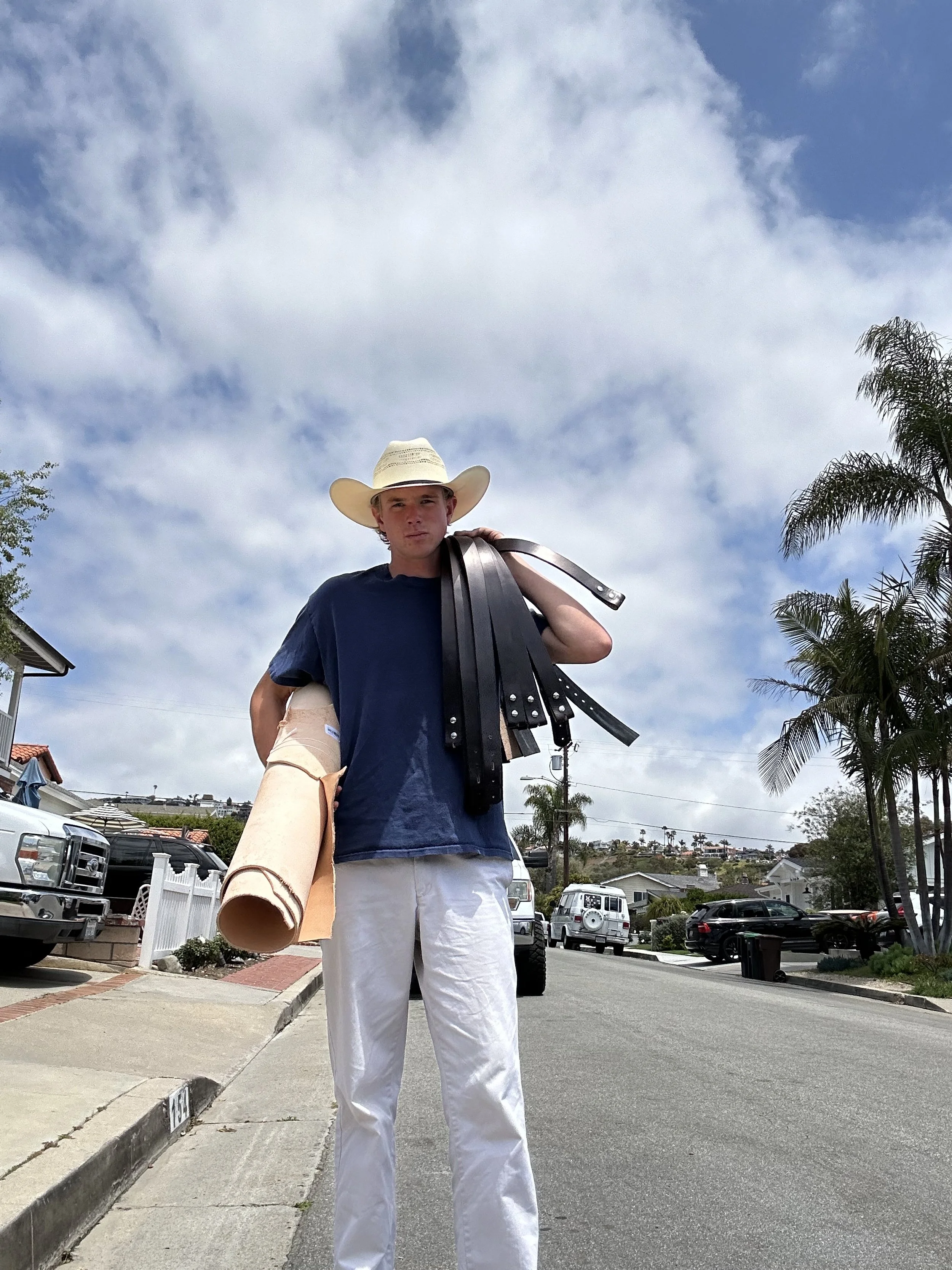 A young man wearing a cowboy hat, a navy Blue t-shirt, and white pants stands on the street holding several black belts and a rolled-up leather hide. Palm trees and parked cars are visible in the background.