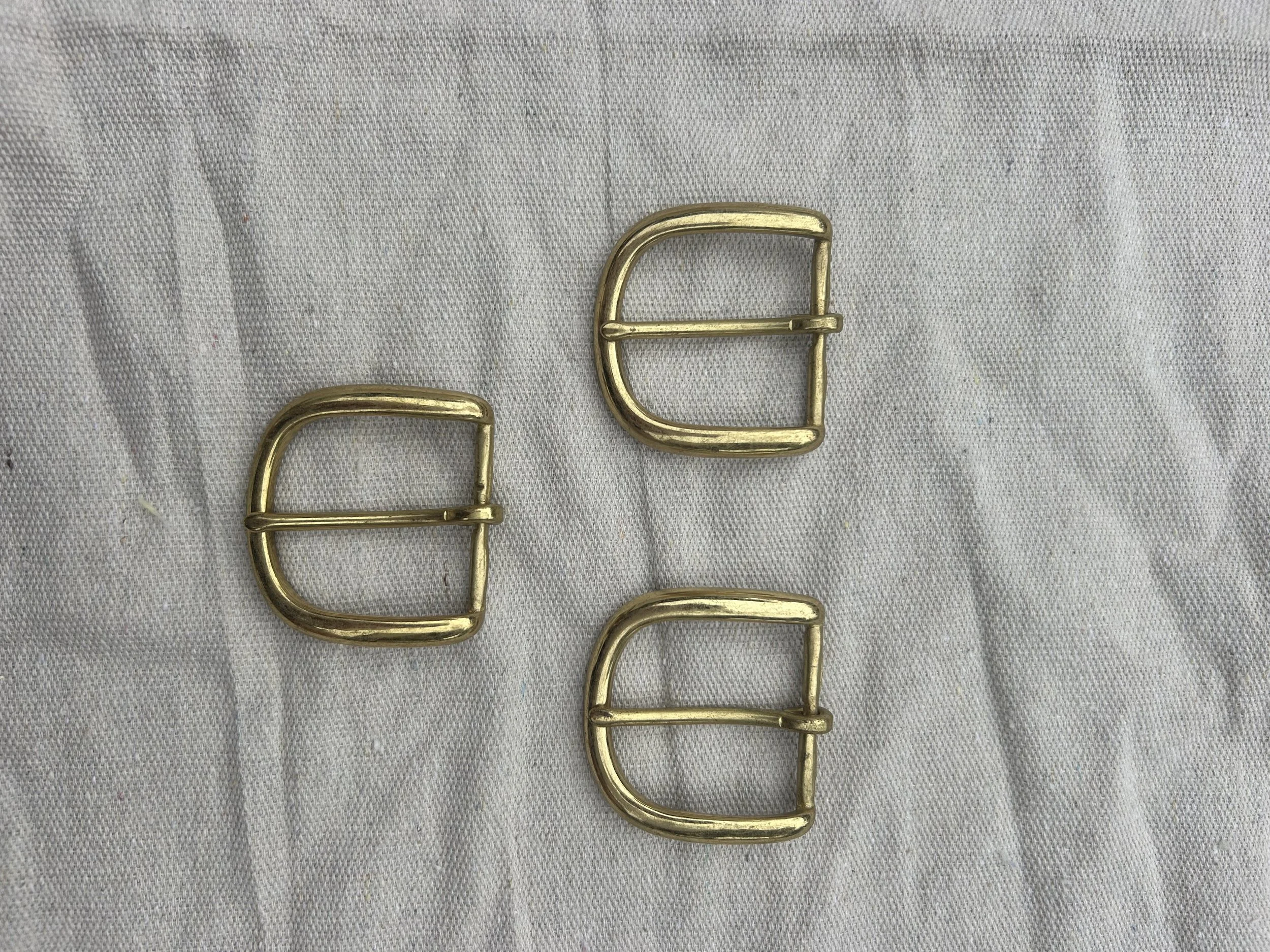 Three gold-colored rectangular belt buckles with rounded edges and a central bar, placed on a light-colored fabric background.