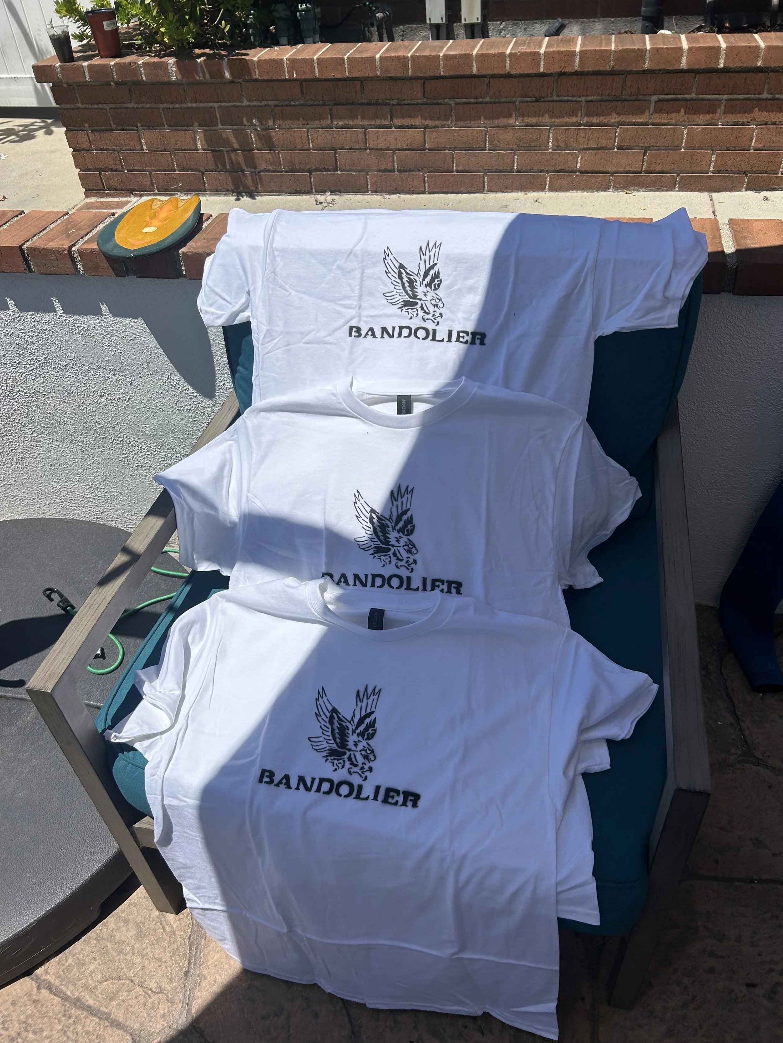 Three white T-shirts with a black eagle logo and the word 'BANDOLIER' printed on them, laid out on a teal cushion on an outdoor chair.