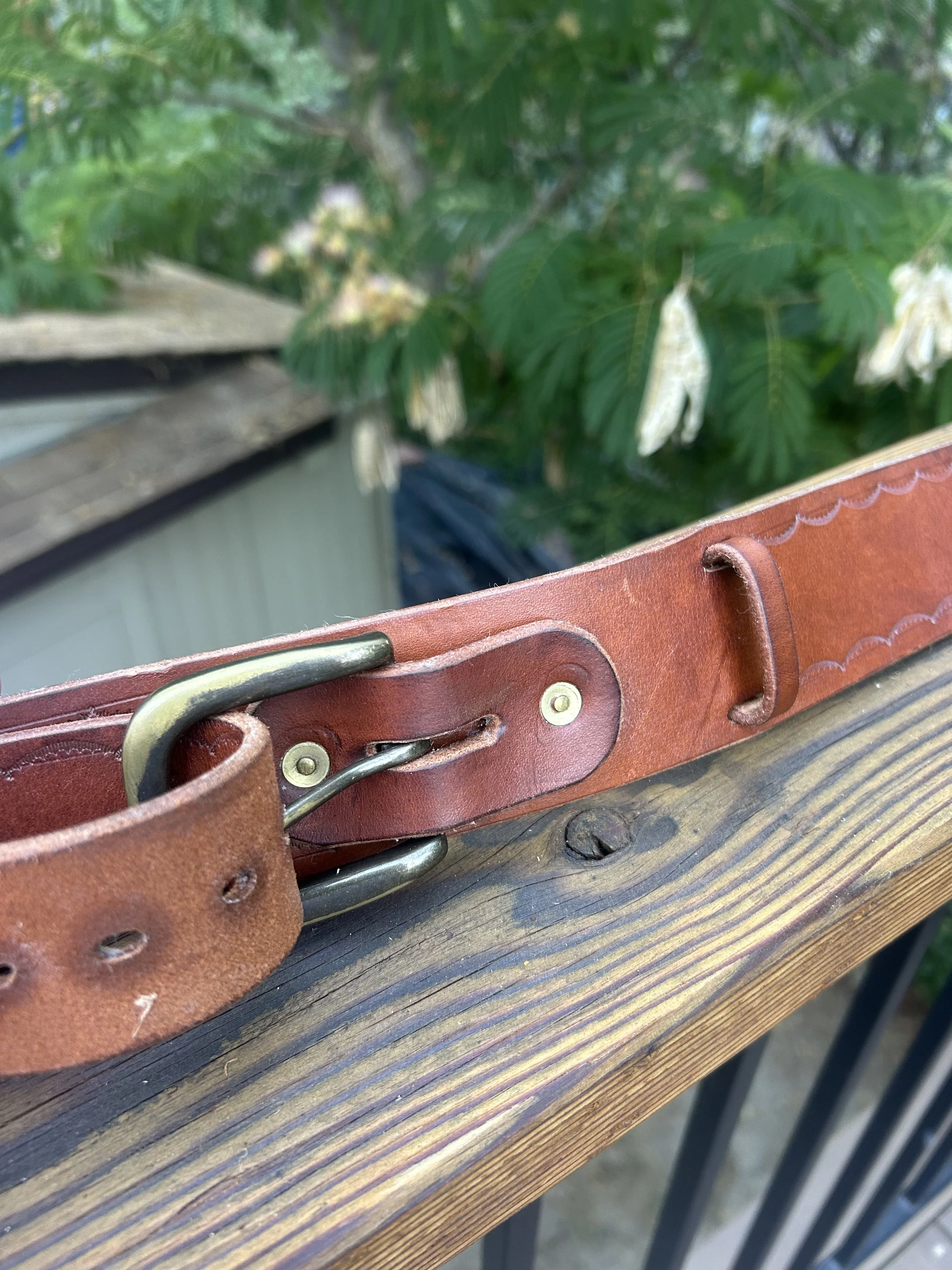 Close-up of a worn brown leather belt with brass buckle resting on a wooden surface, with a background of green leaves and white flowers.