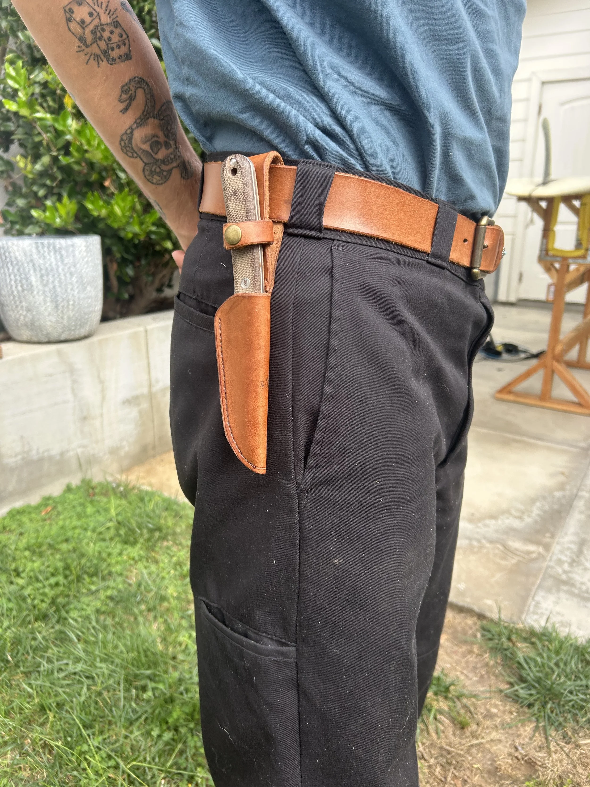 A person standing outdoors with a gun tucked into their leather belt, which is fastened with a brass buckle, and wearing black pants and a blue shirt. The person's arm has tattoos, including a snake tattoo and dice tattoos.